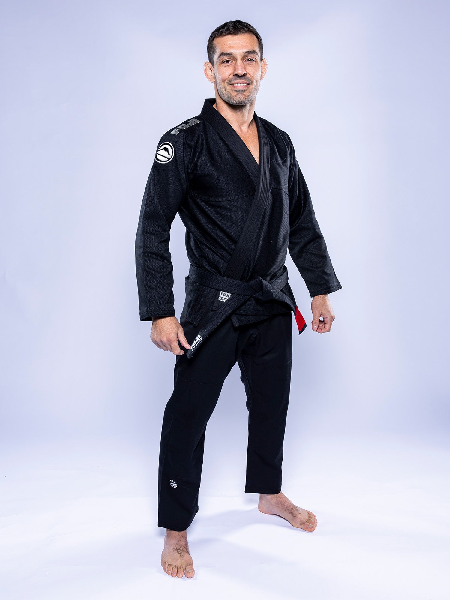 On a white background, a man smiles barefoot with hands at his sides, wearing the Baseline 2.0 BJJ Gi Black Grey and a black belt.
