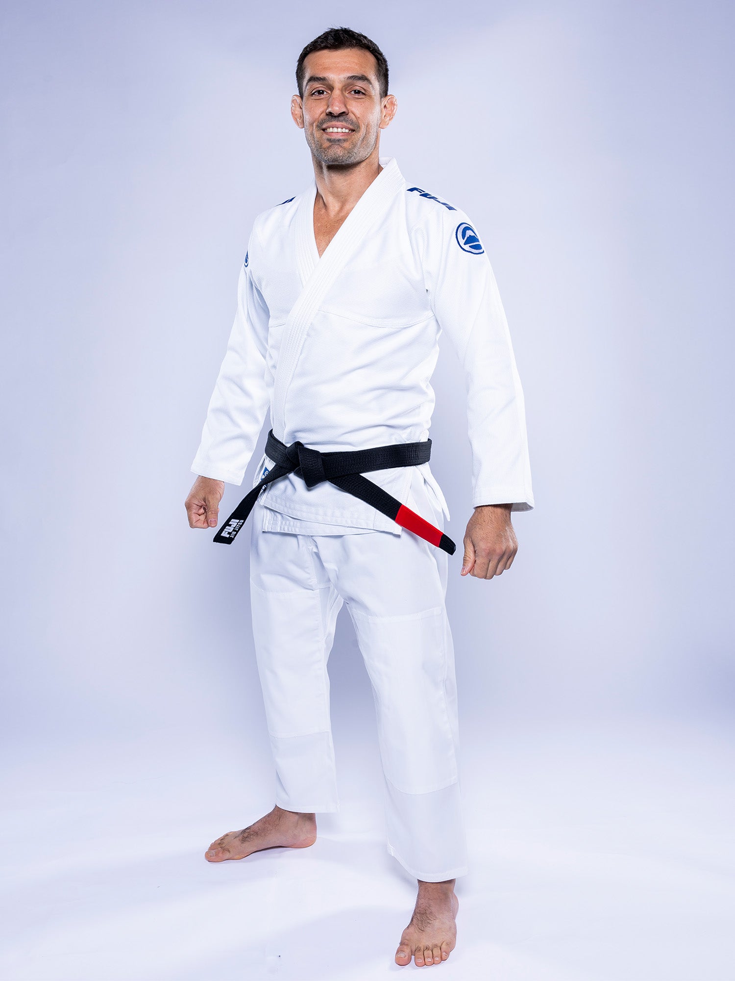 A man in the Baseline 2.0 BJJ Gi White Navy with a black belt stands barefoot on a white background, smiling and facing the camera.
