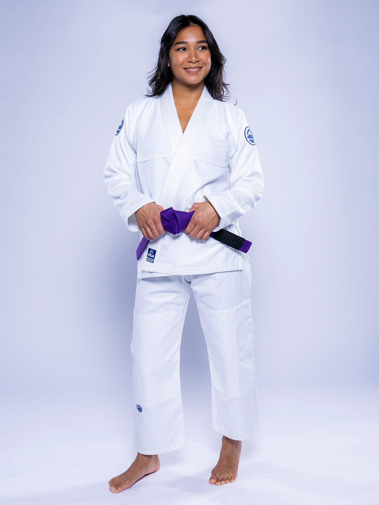 A woman wears the Womens Baseline 2.0 BJJ Gi in white/navy with a purple belt, standing barefoot on a plain white background, smiling and holding her belt with both hands.
