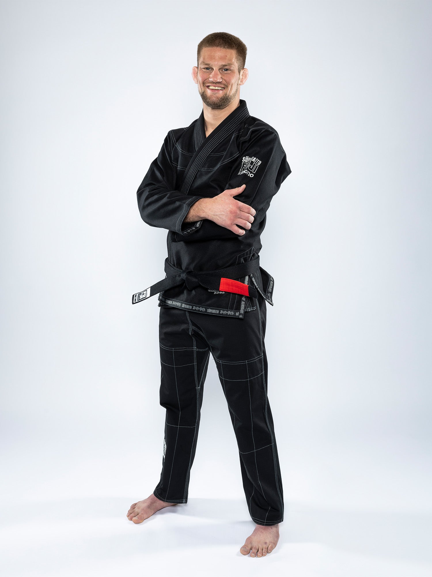 A man stands barefoot on a white background, smiling with his arms crossed while wearing the Suparaito BJJ Gi Black and a red and black belt.