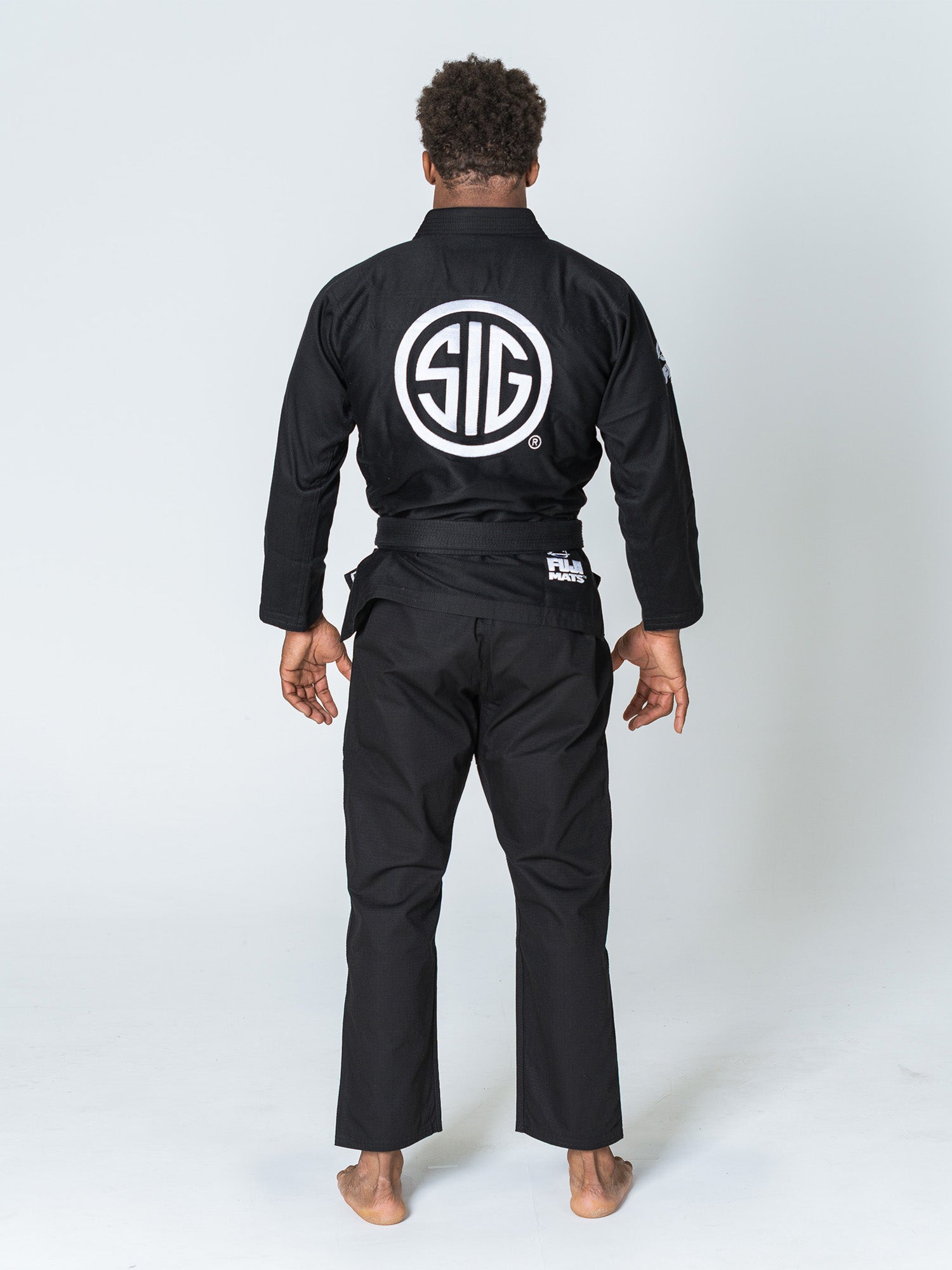 A person stands barefoot on a plain white background, facing away and wearing the Sig Sauer Suparaito Gi Black, which features a large white circular SIG logo on the back.