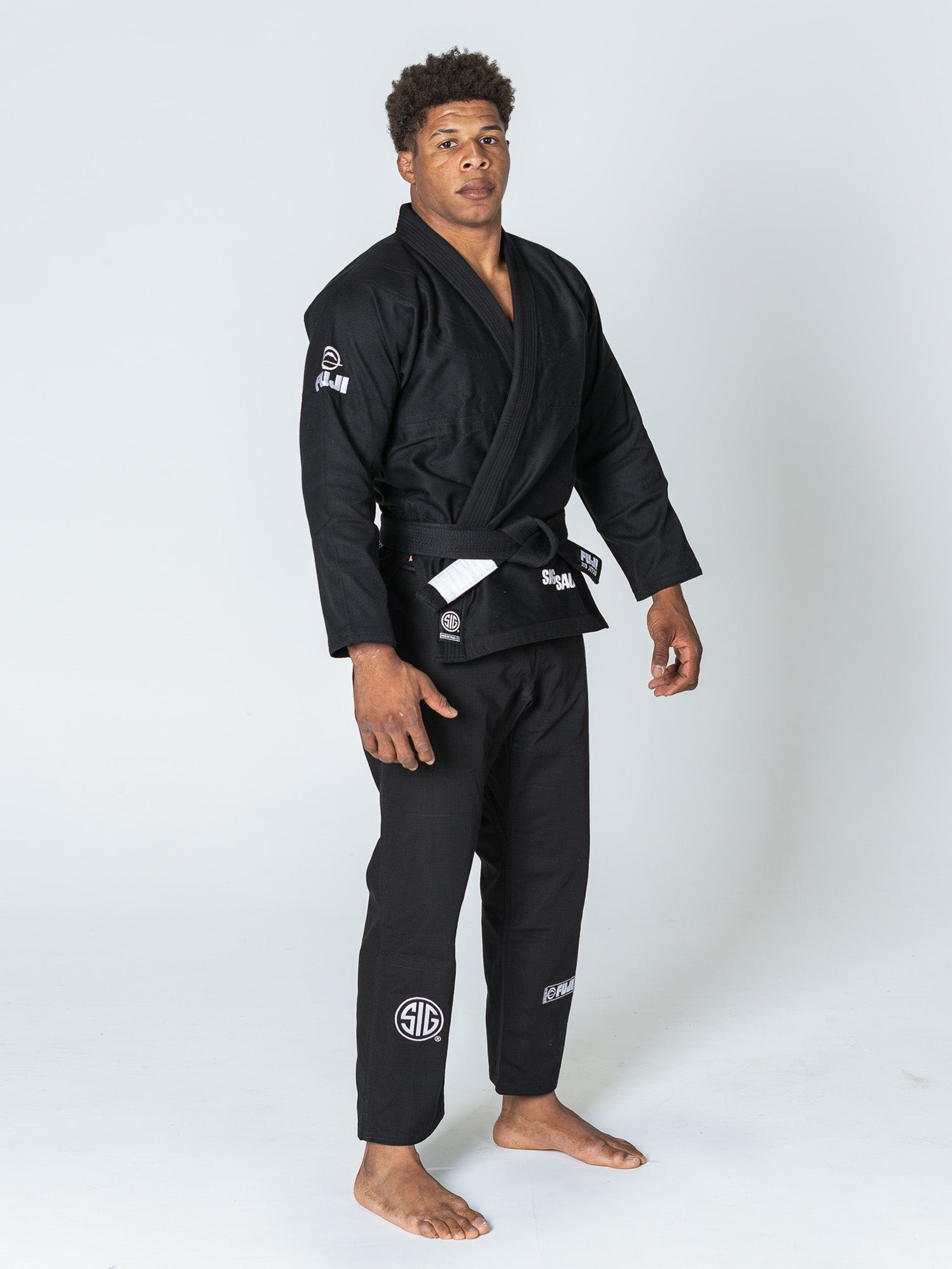 A barefoot man stands on a plain white background wearing a SIG SAUER Suparaito Gi Black with several white patches and a black belt.