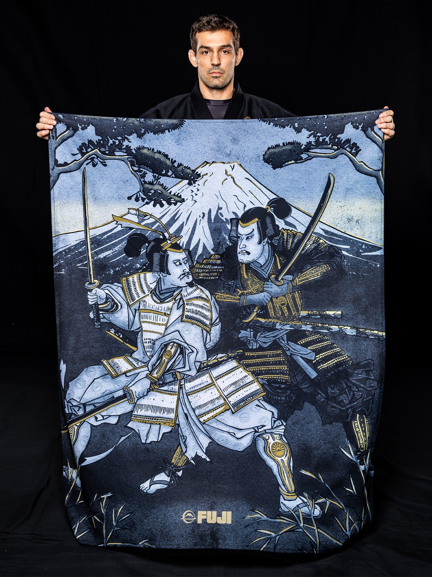 A man holds a Heritage Keishō Edition Bundle blanket featuring artwork of two samurai in armor with swords drawn, standing under a tree with Mount Fuji in the background.