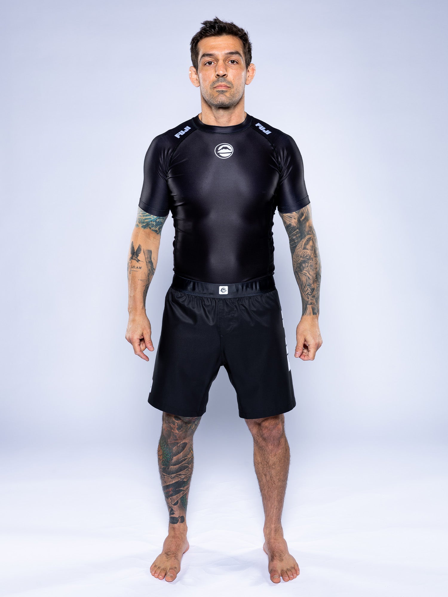 A man with tattoos on both arms and his left leg stands barefoot against a plain backdrop, facing forward in a neutral pose. He wears black Competition Ranked Shorts and a black short-sleeved rash guard.