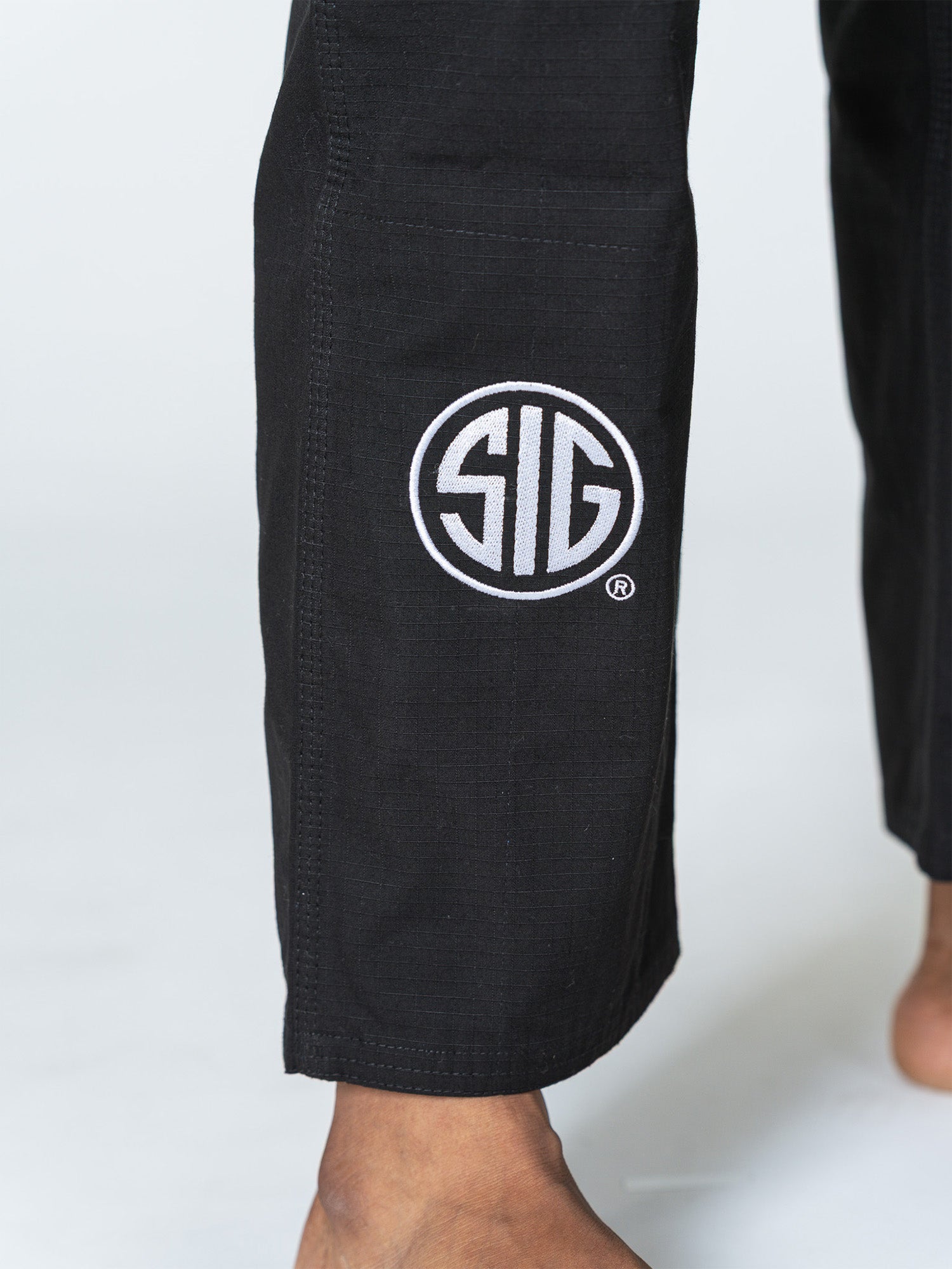 Close-up of a person’s lower legs barefoot, wearing black Sig Sauer Suparaito Gi pants featuring a white circular SIG logo near the left ankle, standing on a white surface.