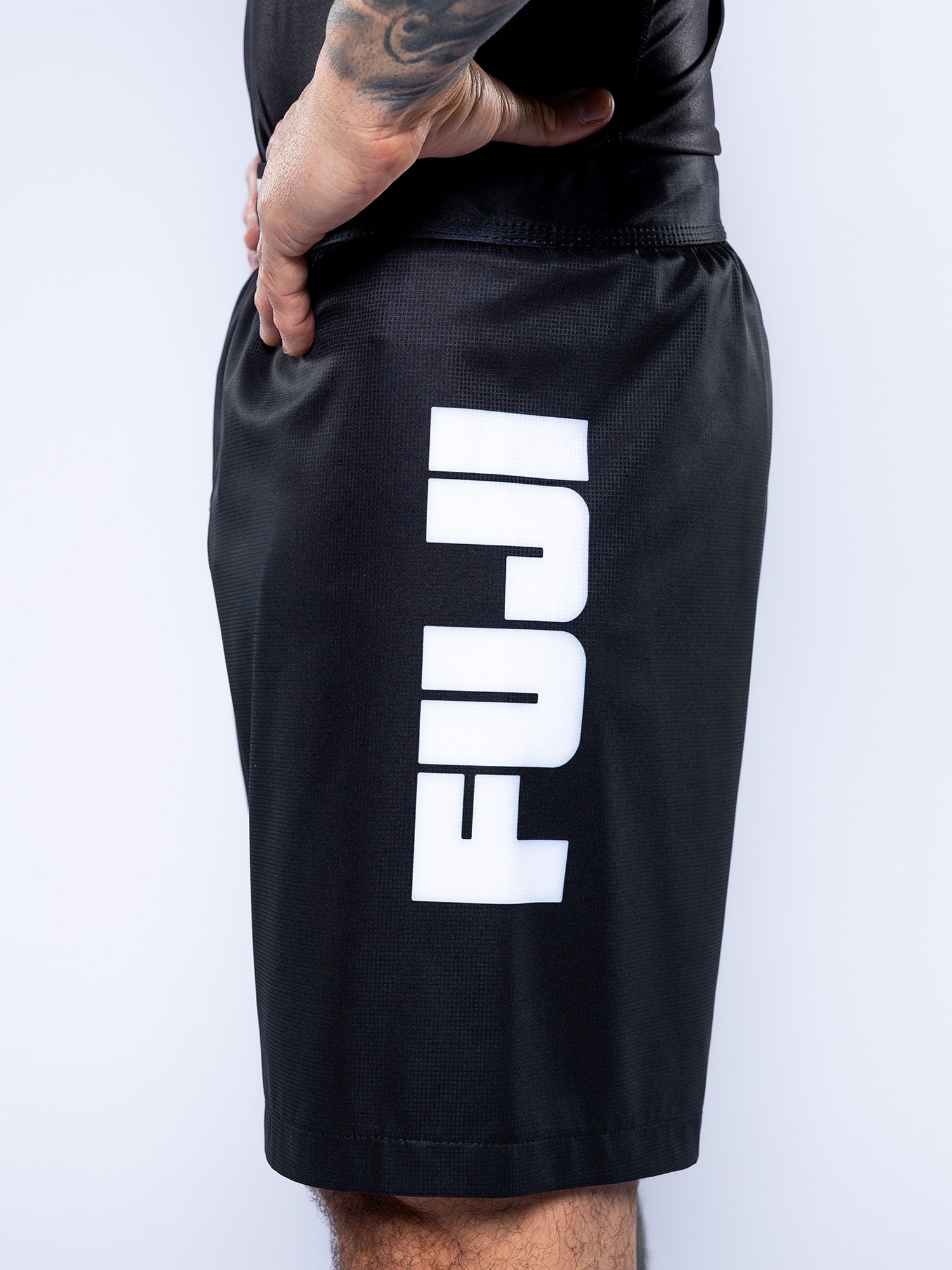 Someone wearing Competition Ranked Shorts Black, featuring "FUJI" in bold white letters down the left leg. Their hand is on their hip and a forearm tattoo is visible.