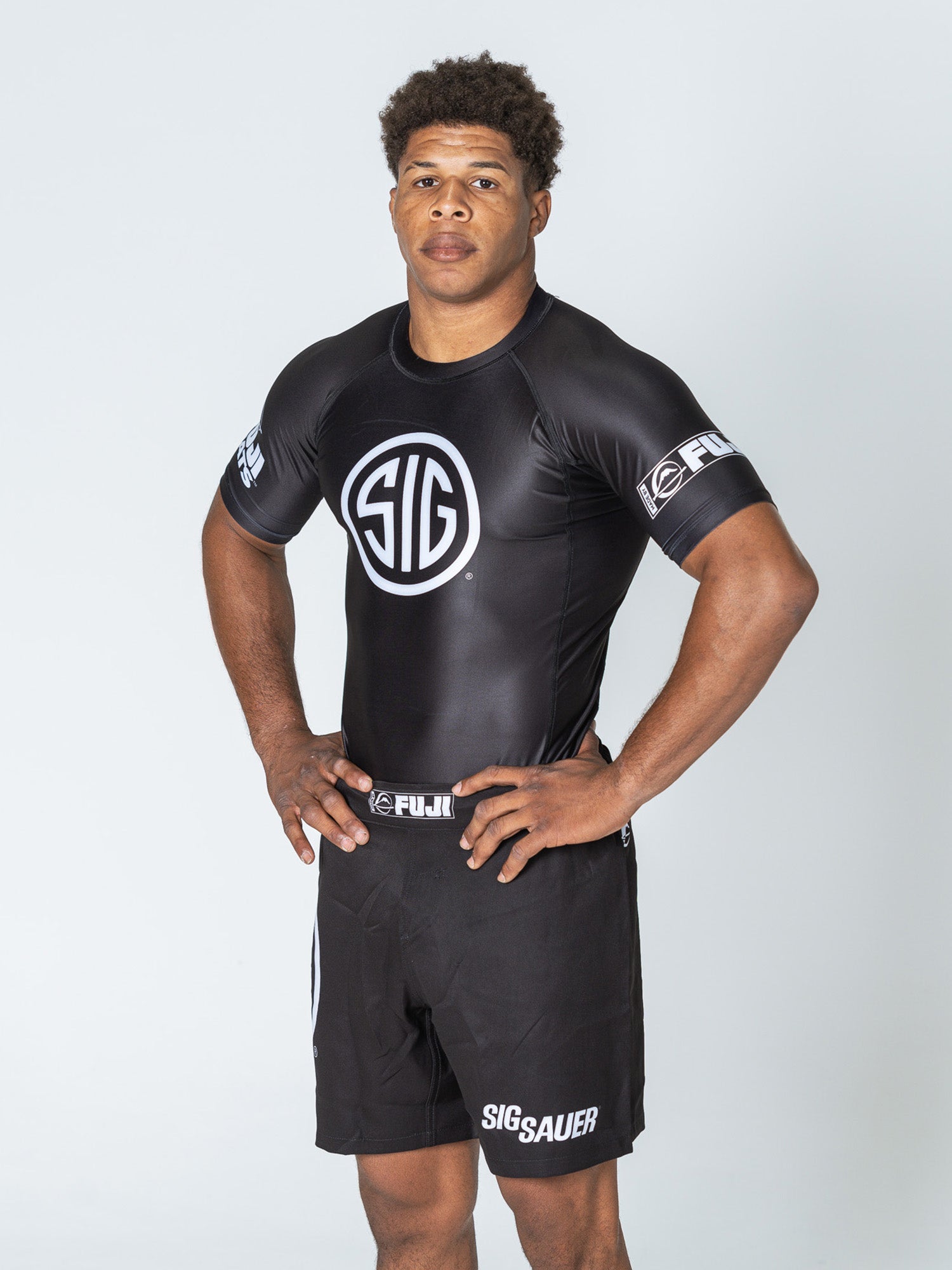 A man stands with hands on hips wearing the Sig Sauer Short Sleeve Rashguard in black and matching shorts with white SIG and Sig Sauer logos, posing against a plain light background.