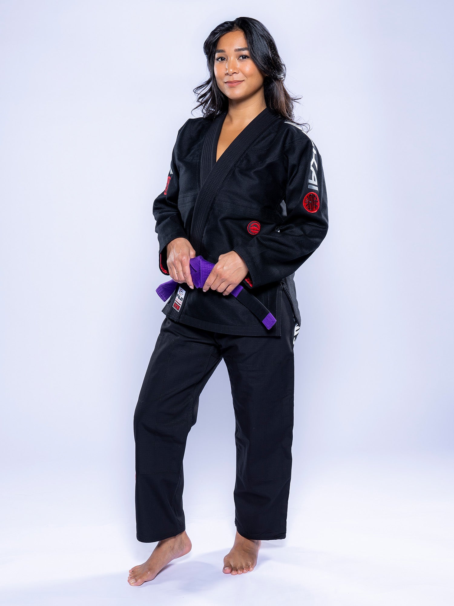A woman wearing the Womens SEKAI 3.0 BJJ Gi Black Grey holds a purple belt in front of her, barefoot on a white background, looking at the camera with a slight smile.