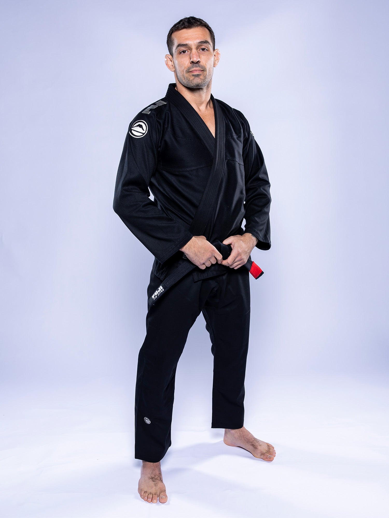 A man in a Baseline 2.0 BJJ Gi Black Grey stands barefoot and confident on a white background, holding his black belt with red bar in front of him.