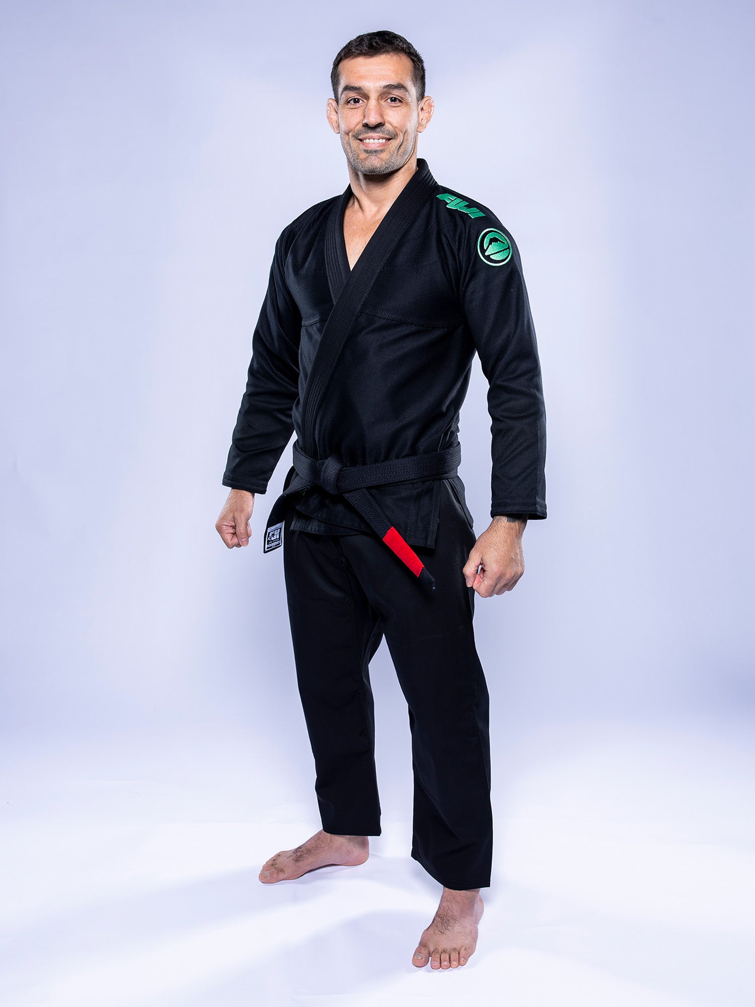 A man wearing the Baseline 2.0 BJJ Gi Black Mint and a black belt stands barefoot on a white background, smiling at the camera.
