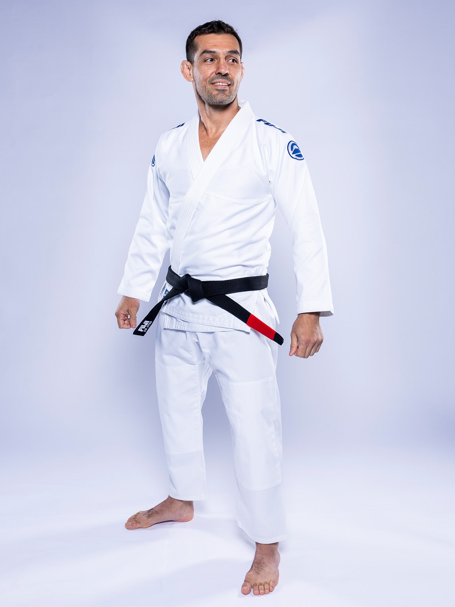 A man wearing the Baseline 2.0 BJJ Gi in white/navy with a black belt stands barefoot on a white background, smiling and looking slightly to the side.