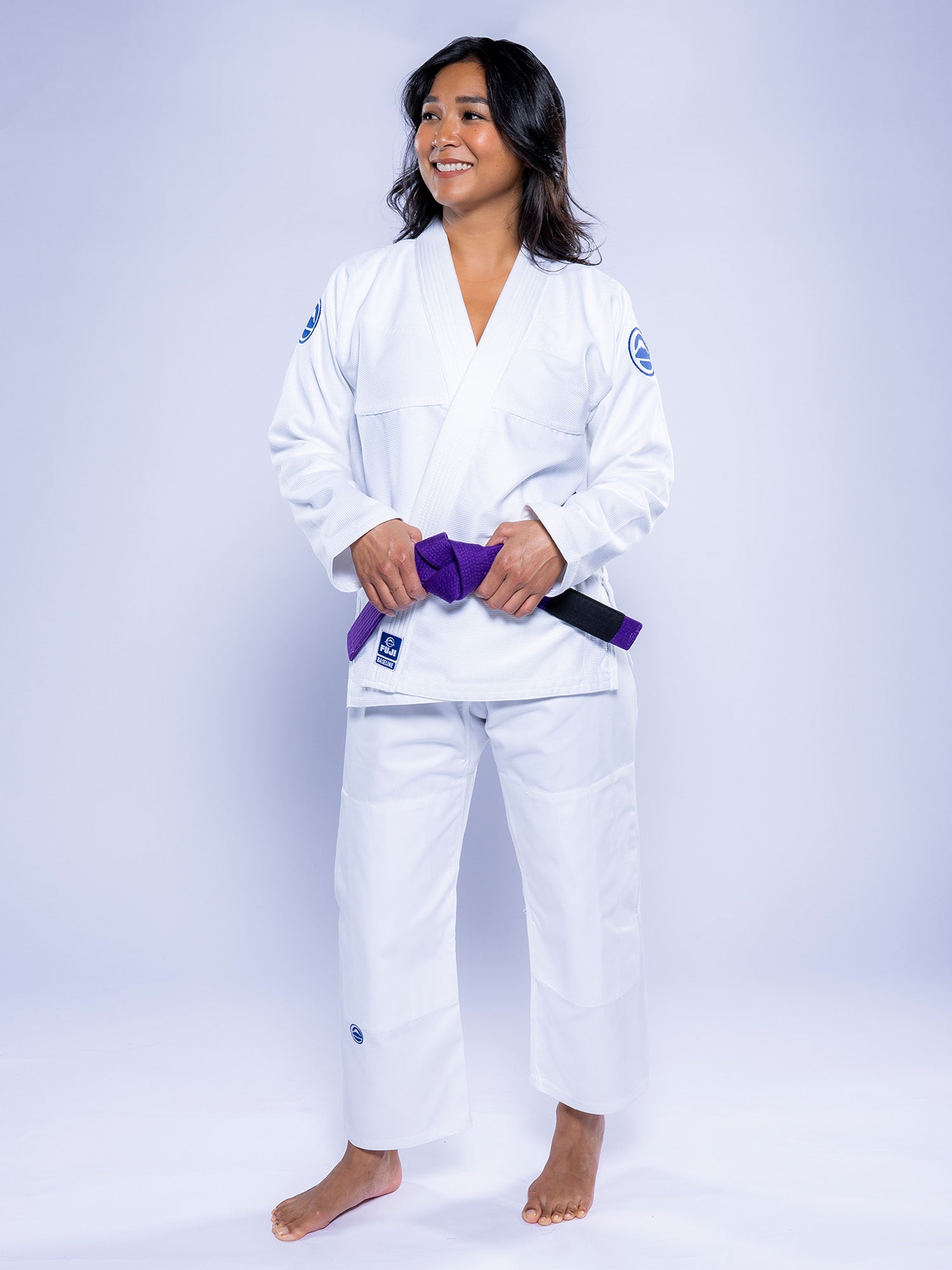 A woman wearing the Womens Baseline 2.0 BJJ Gi in white with navy accents and a purple belt stands barefoot, smiling and looking to the side against a plain light background.