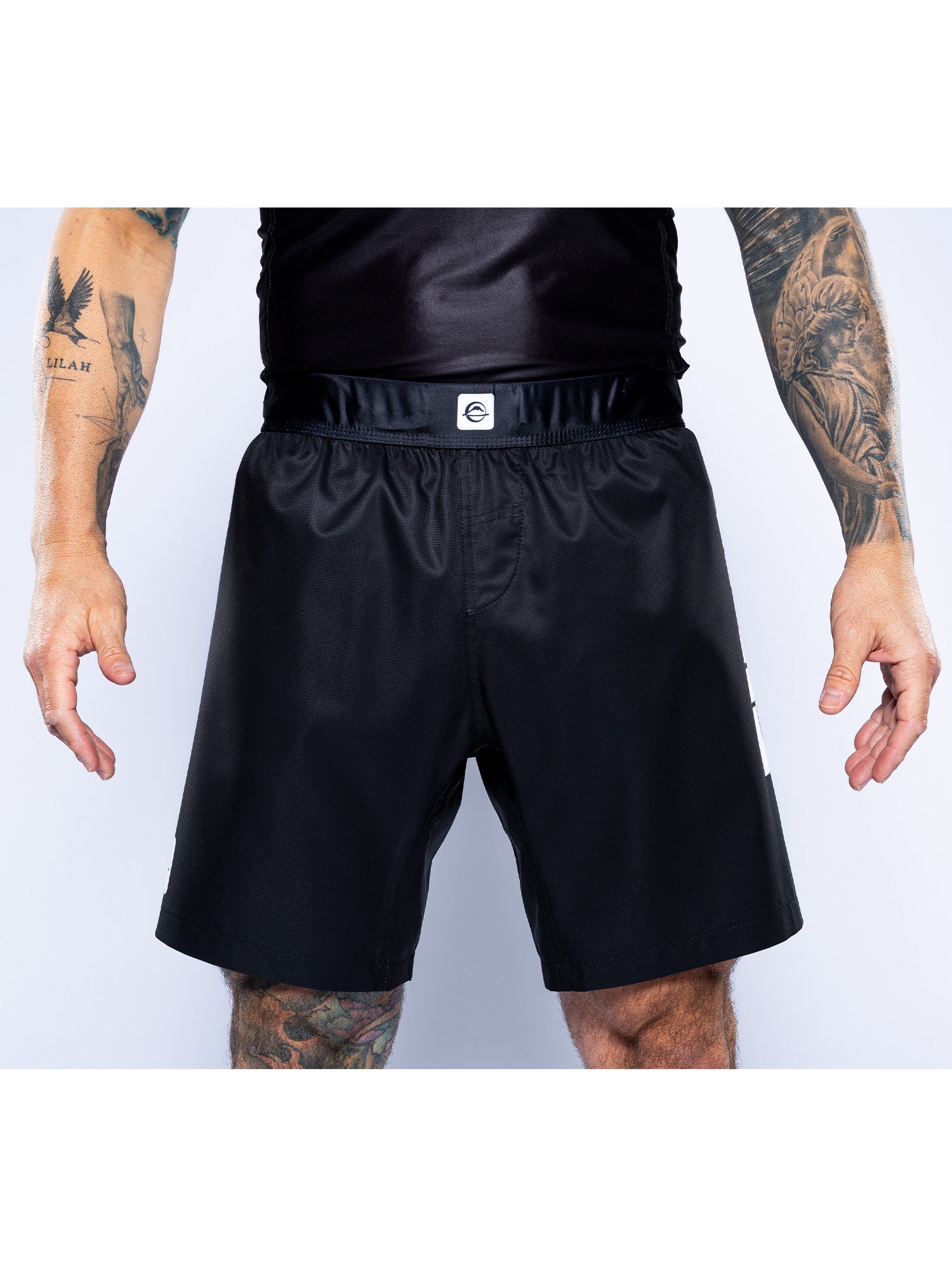 A person models Competition Ranked Shorts Black, paired with a black shirt, standing against a plain light background with tattooed arms and legs visible.