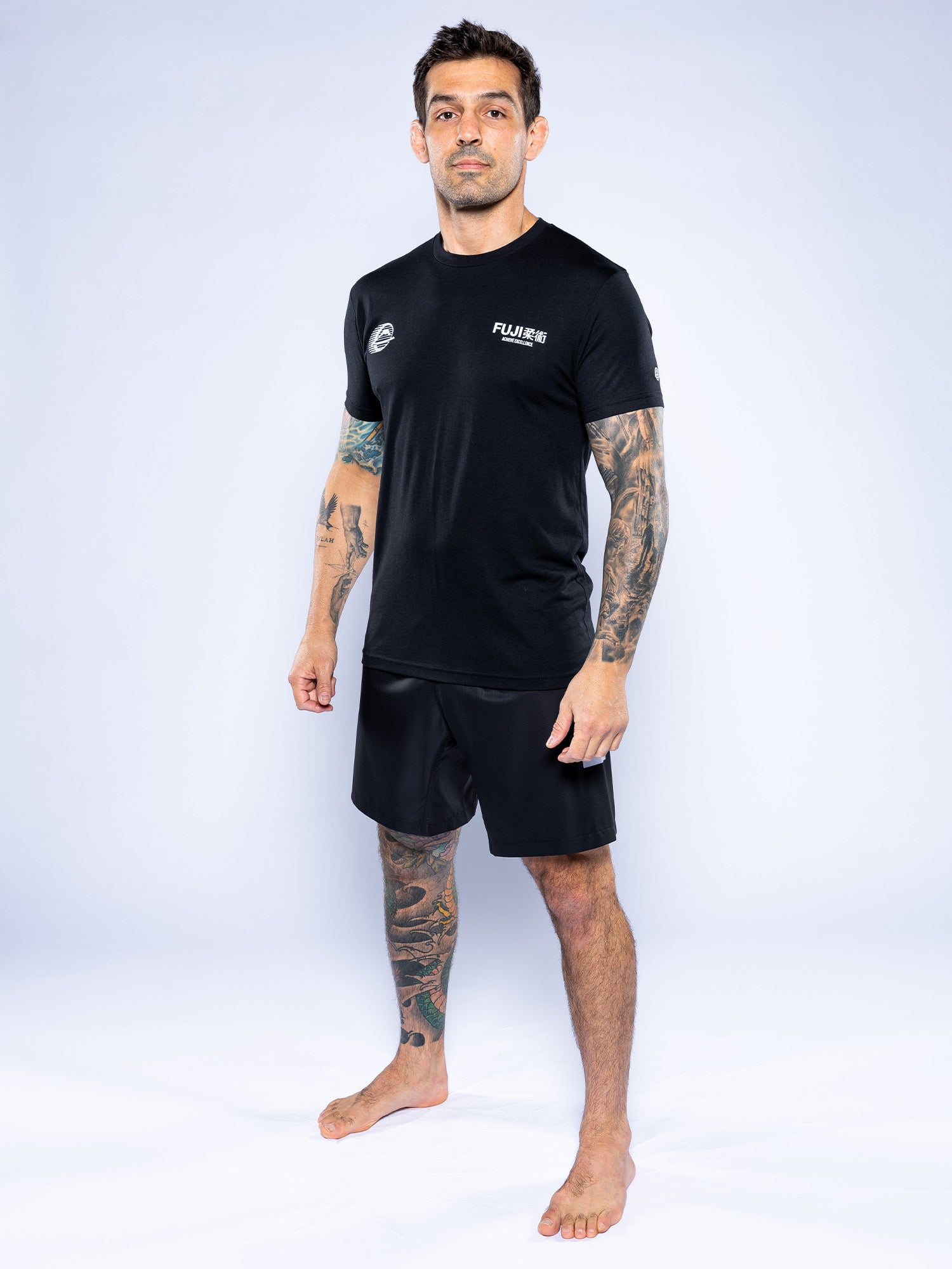 A tattooed man stands barefoot on a white background, confidently wearing the Iconic Jiu Jitsu Shirt Black and black shorts, looking at the camera.