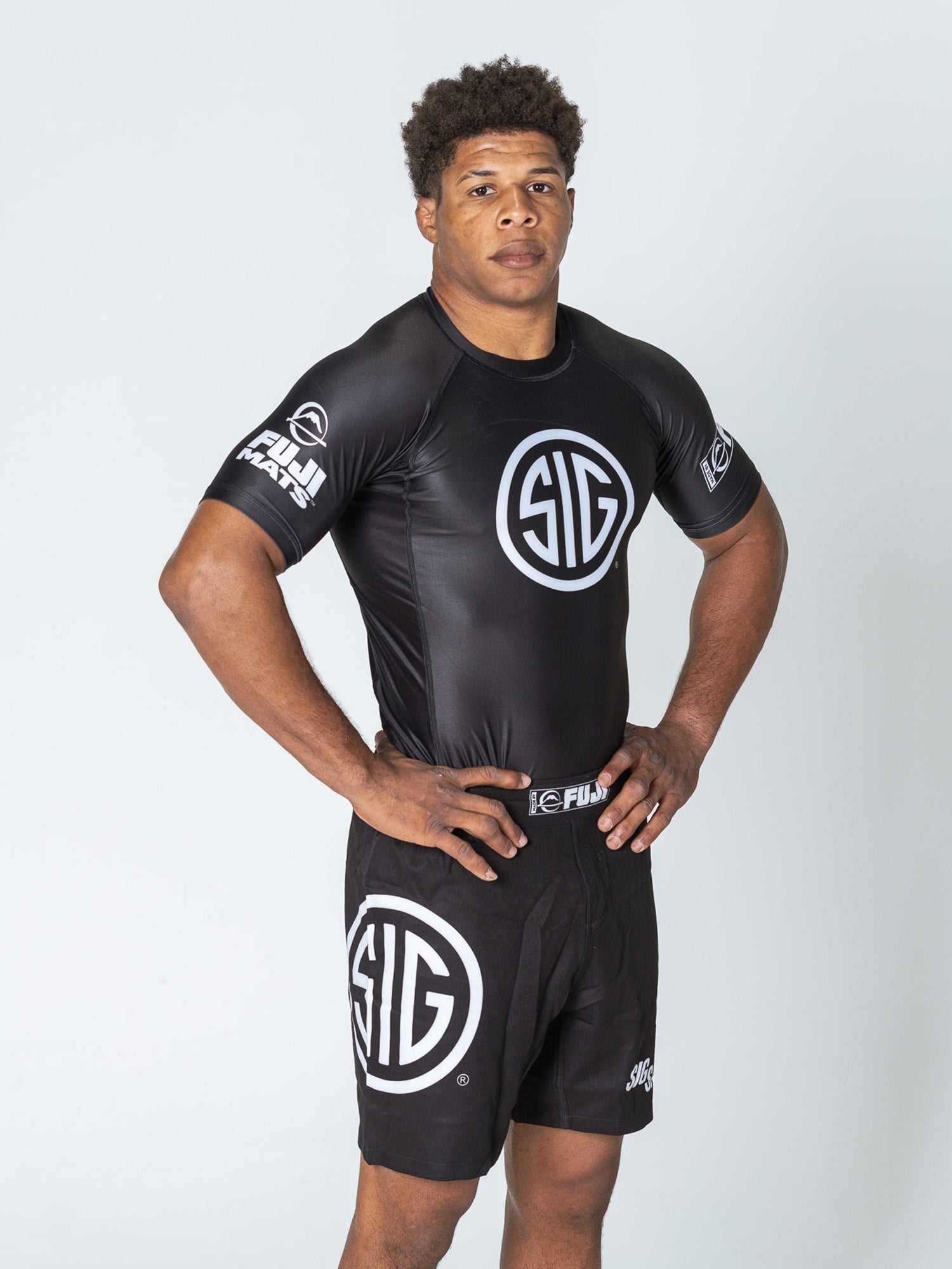 A man with short curly hair stands against a plain background, wearing the Sig Sauer Short Sleeve Rashguard in black and matching shorts with white SIG logos. He has one hand on his hip and a neutral expression.