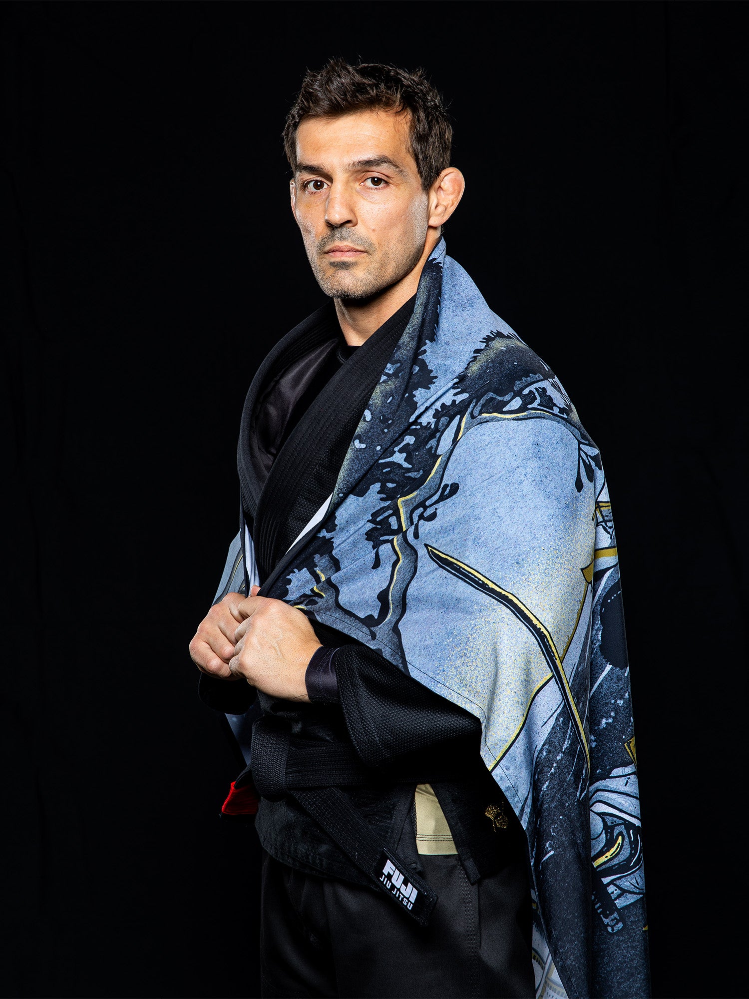 A man wearing the Heritage Keisho Lightweight BJJ Gi in black stands against a black background, draping a blue, illustrated cape or blanket over his shoulders while looking at the camera.