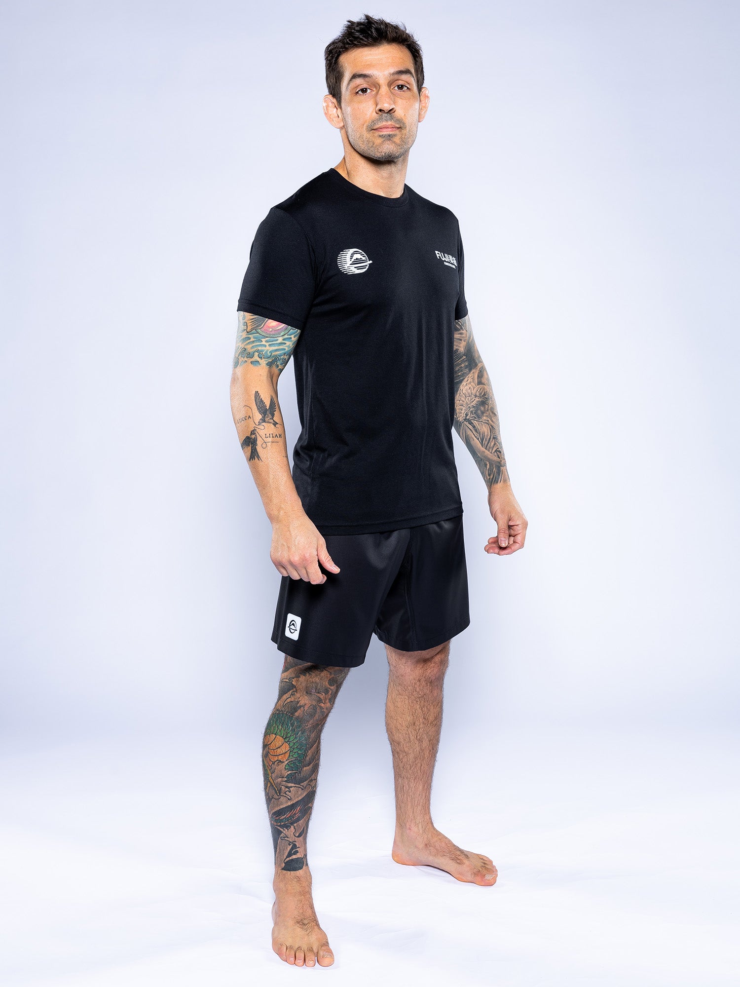 A tattooed man stands barefoot on a white background, wearing the Iconic Jiu Jitsu Shirt Black and black athletic shorts, looking at the camera with a neutral expression.
