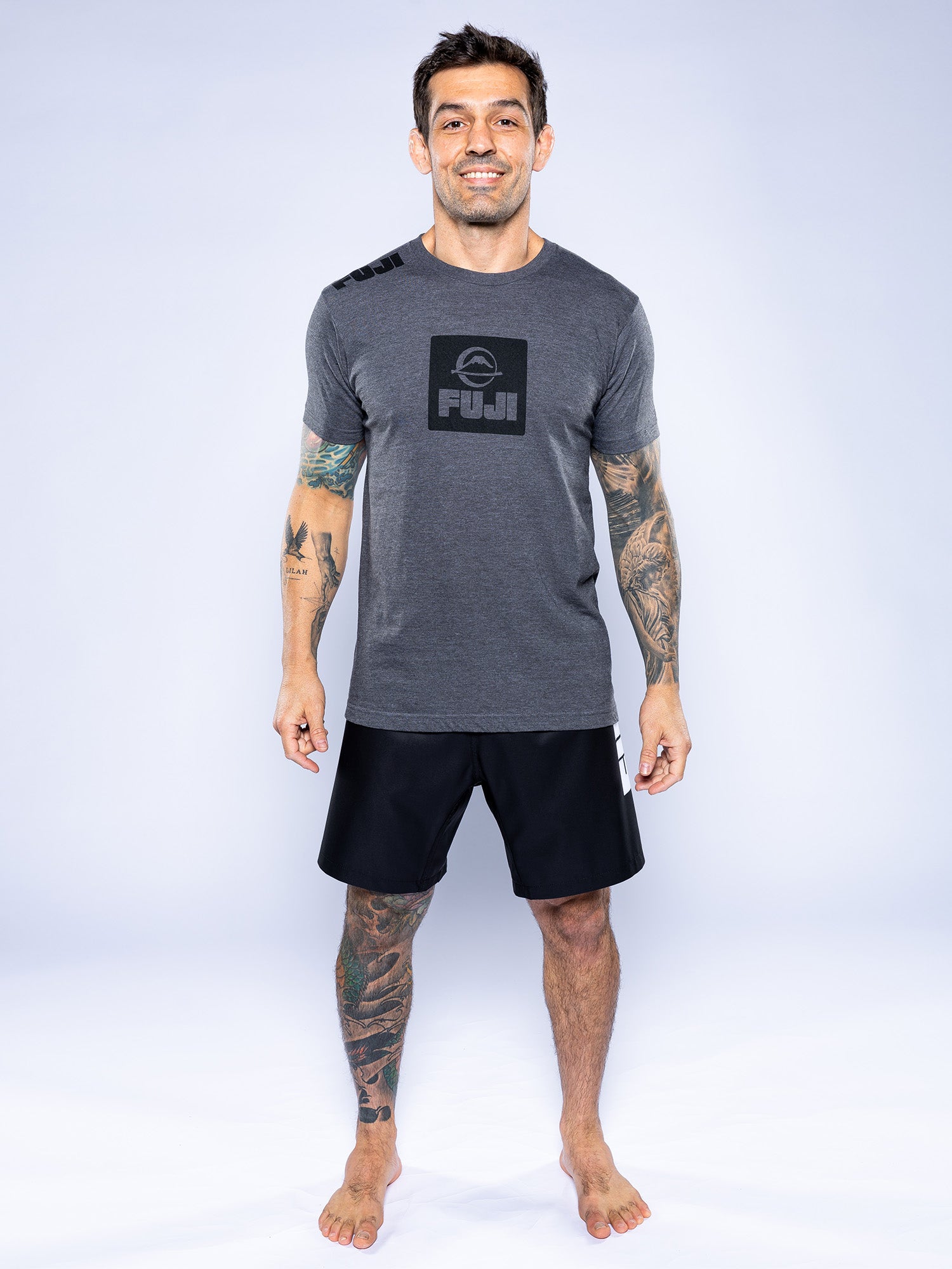 A man with tattoos on his arms and legs stands barefoot, smiling, wearing a charcoal Elevate Your Journey Shirt and black shorts against a light background.