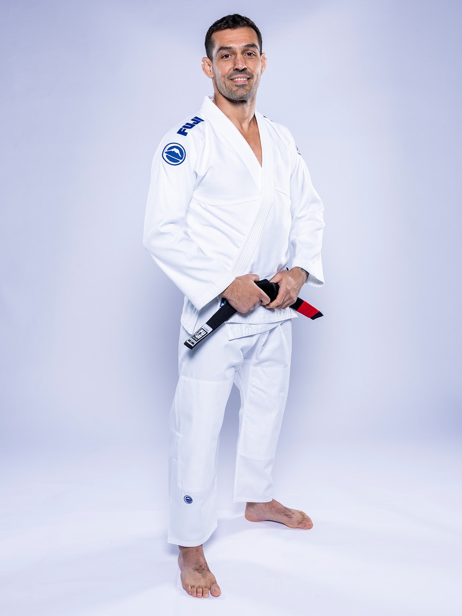 A man stands barefoot and smiling in front of a plain light background, wearing the Baseline 2.0 BJJ Gi White Navy and holding a black belt with a red bar.