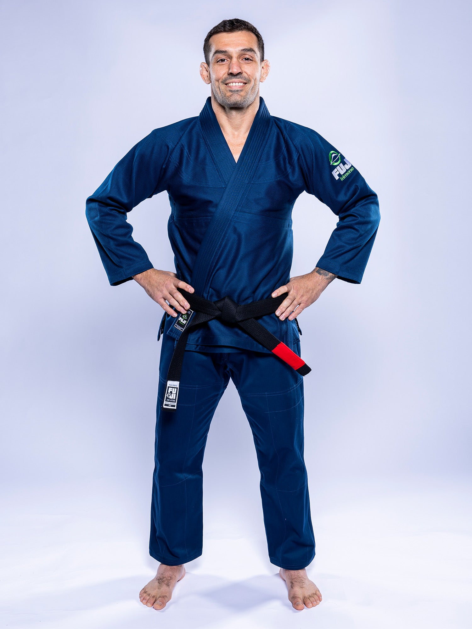 A man stands barefoot on a white background, smiling with his hands on his hips, wearing the Lightweight BJJ Gi Navy and a black belt with a red bar.