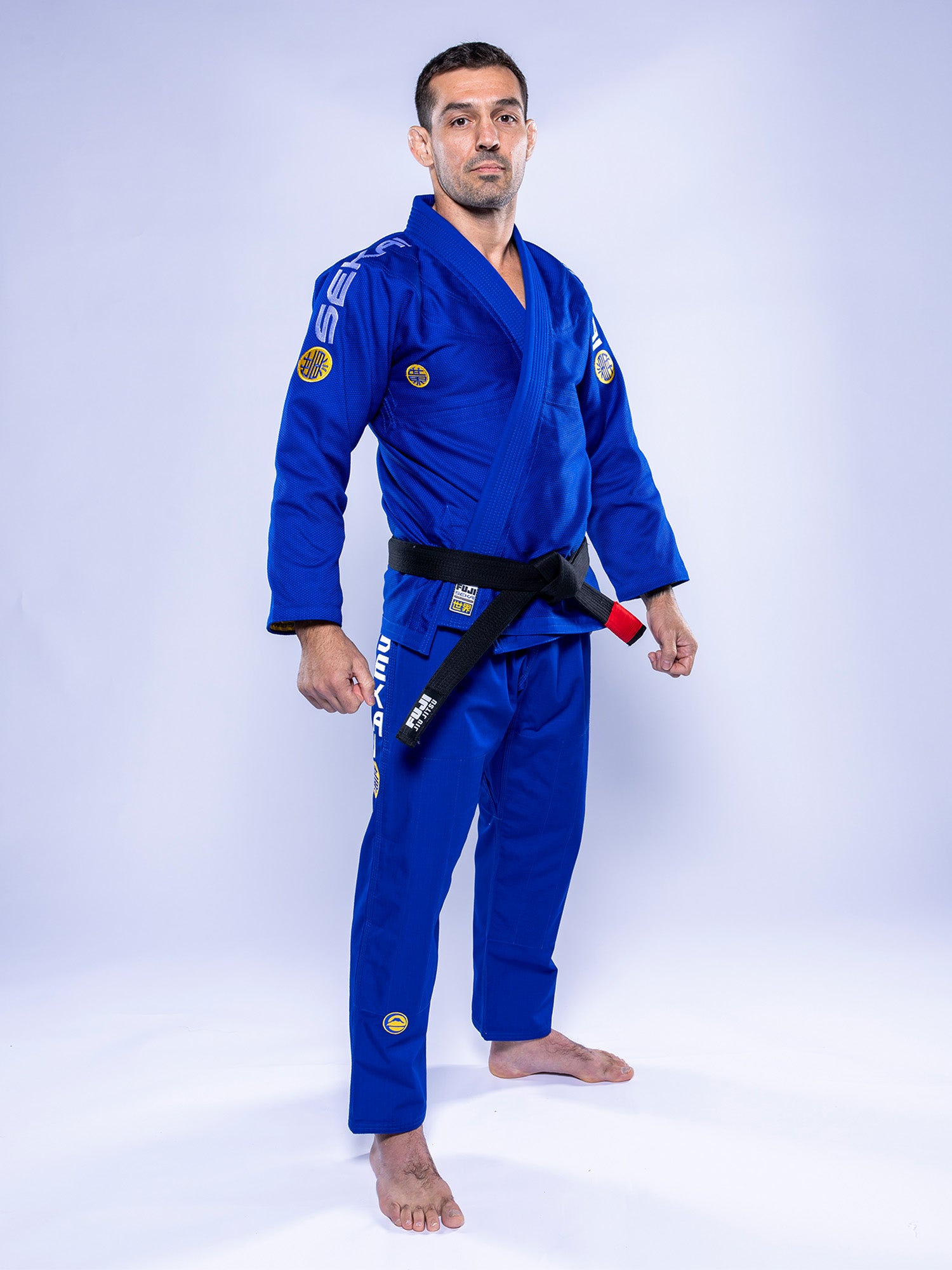 A man wearing the SEKAI 3.0 BJJ Gi Blue Yellow with a black belt stands barefoot on a white background, facing forward with a serious expression.