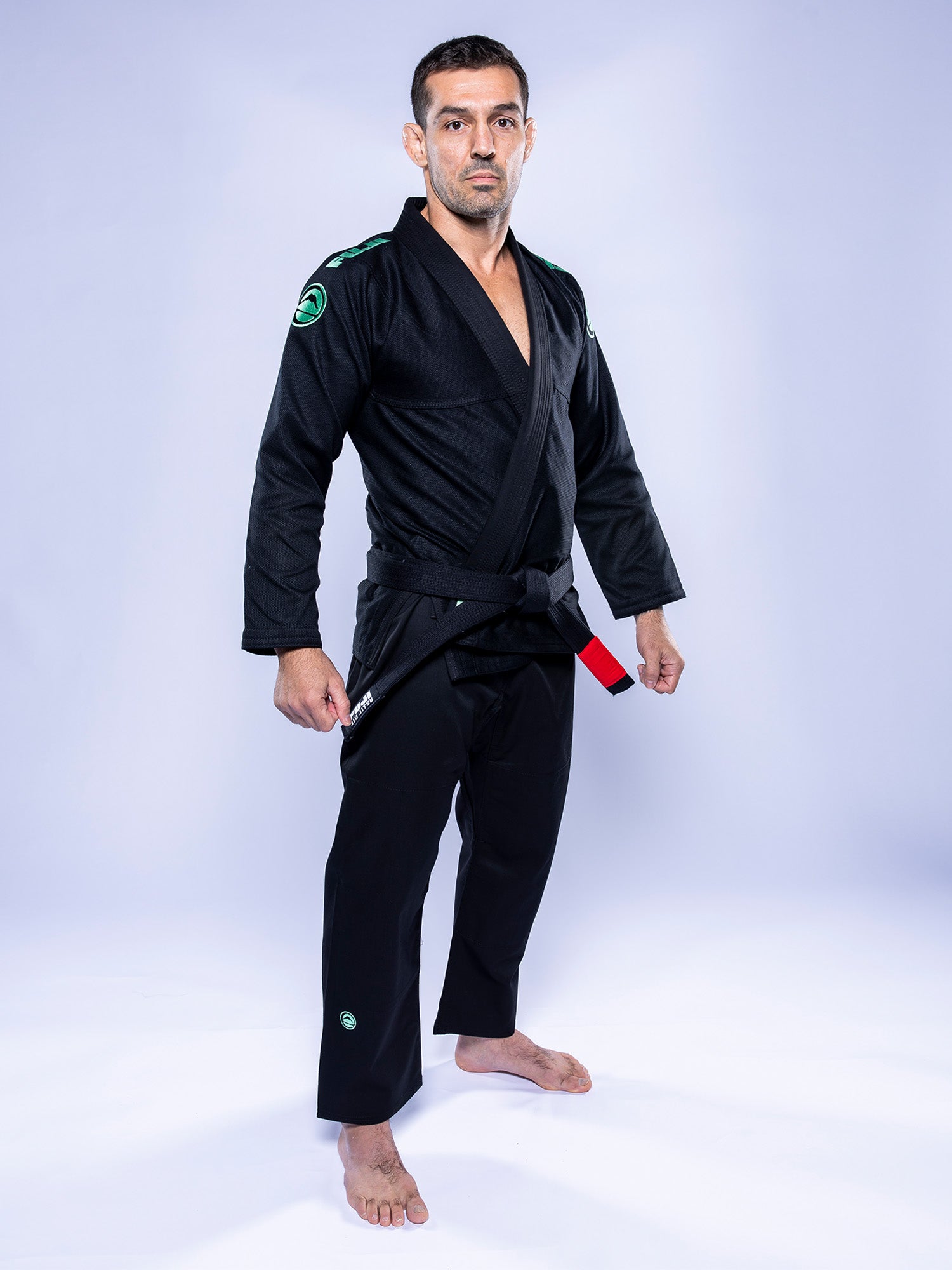A man stands barefoot on a white floor, confidently wearing the Baseline 2.0 BJJ Gi Black Mint and a black belt, looking at the camera against a light background.
