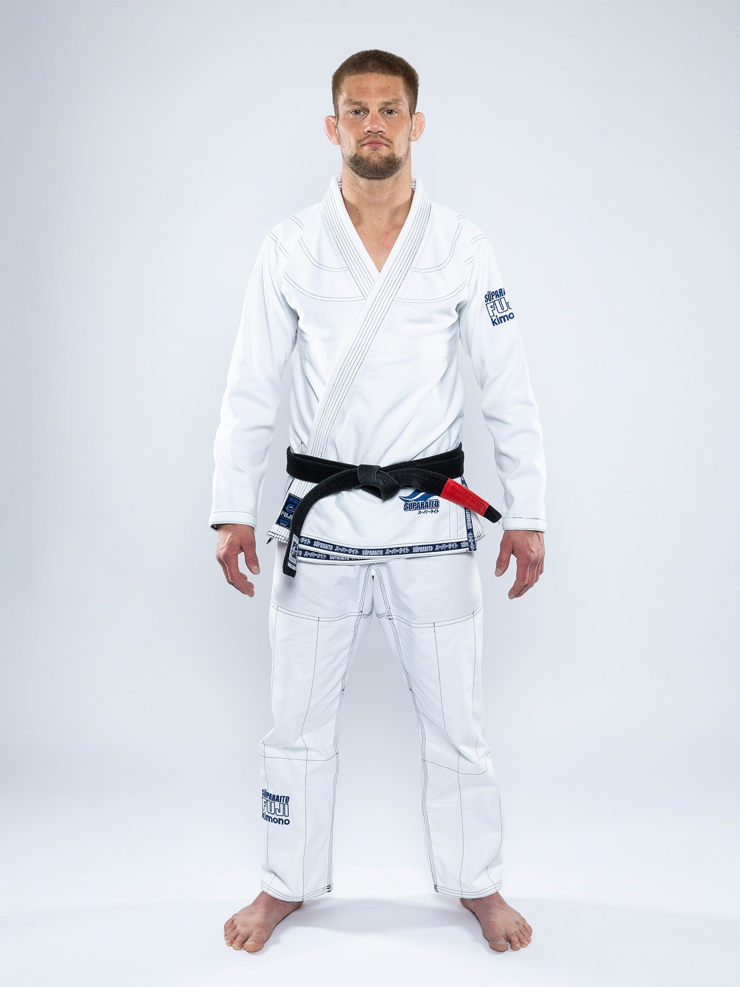 A man with short hair and a trimmed beard stands barefoot on a white floor, wearing the Suparaito BJJ Gi White and a black belt, facing forward with a serious expression against a plain white background.