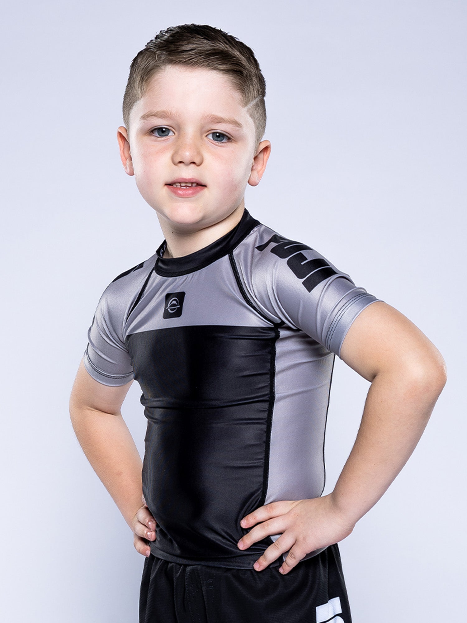 A young boy with short brown hair stands confidently with hands on hips, wearing the Kids Competition Ranked Rashguard Grey and black shorts, posing against a plain light gray background.
