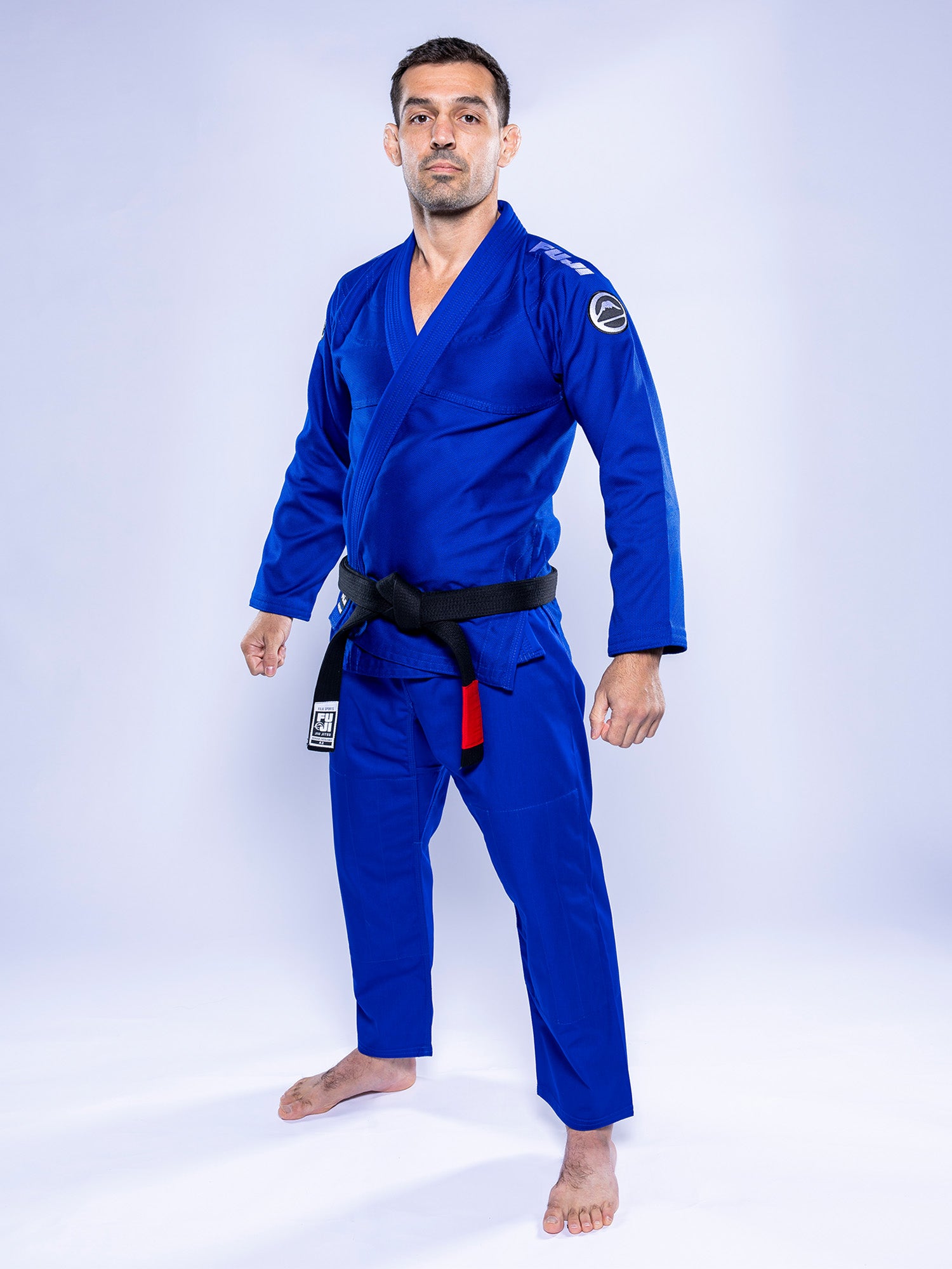 A man stands barefoot on a white background, facing forward with a confident expression, wearing the Baseline 2.0 BJJ Gi Blue White and a black belt, his hands relaxed at his sides.