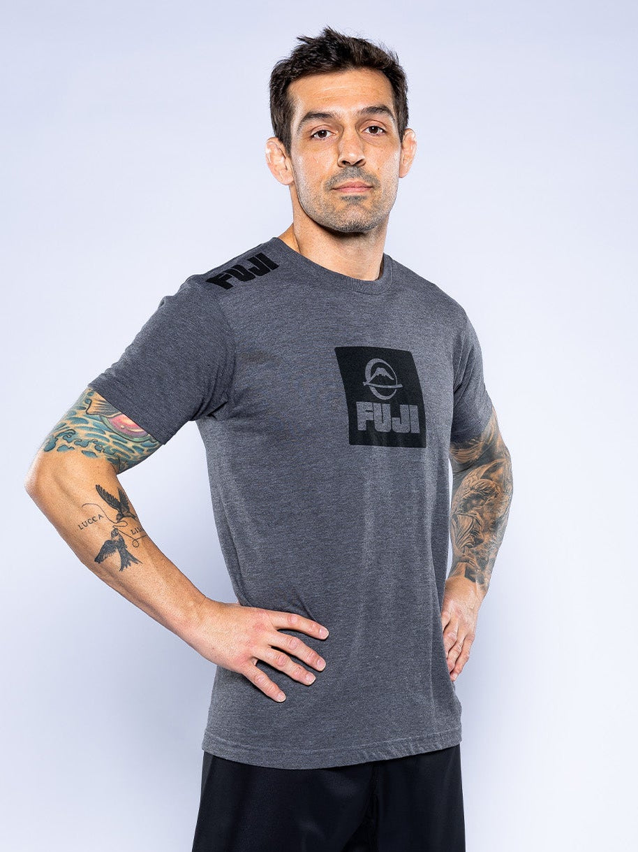 A man with short dark hair and tattoos on both arms and one leg stands barefoot on a white surface, wearing the Elevate Your Journey Shirt Charcoal and black shorts, looking confidently at the camera.