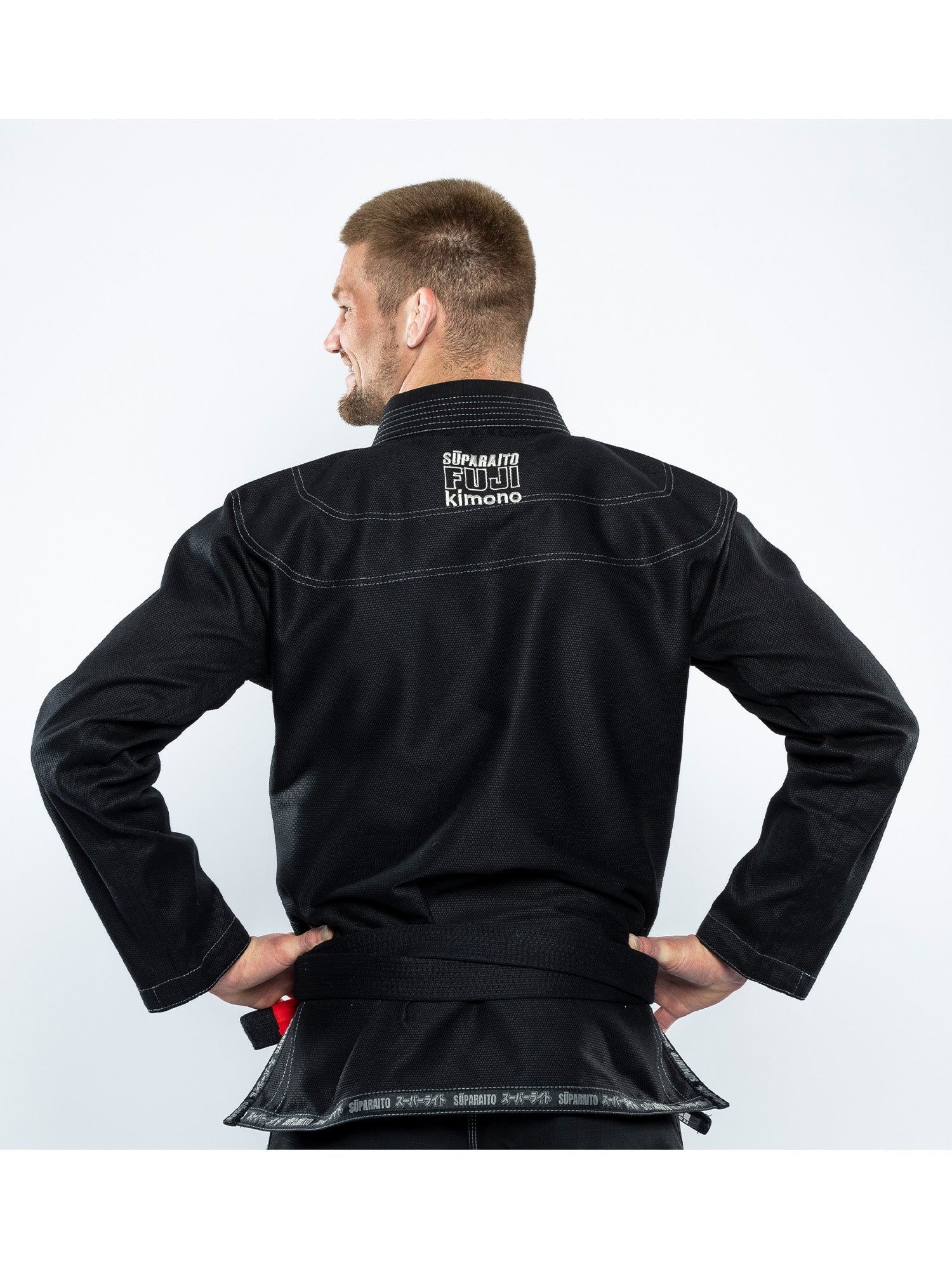 A man stands with his back to the camera wearing the Suparaito BJJ Gi Black, featuring "Shirakawa Fight Kimono" in white lettering on the back collar. His hands are on his hips against a plain white background.