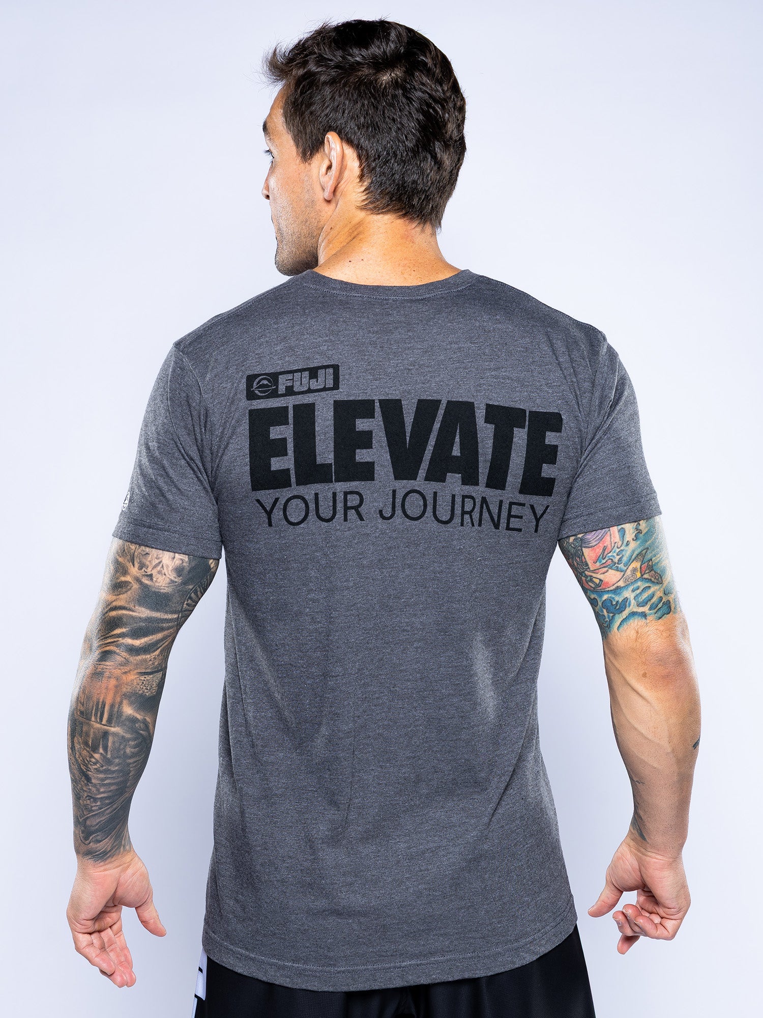 A man with tattooed arms is seen from the back, wearing the Elevate Your Journey Shirt Charcoal featuring "FIJI ELEVATE YOUR JOURNEY" in bold black letters on a plain light background.