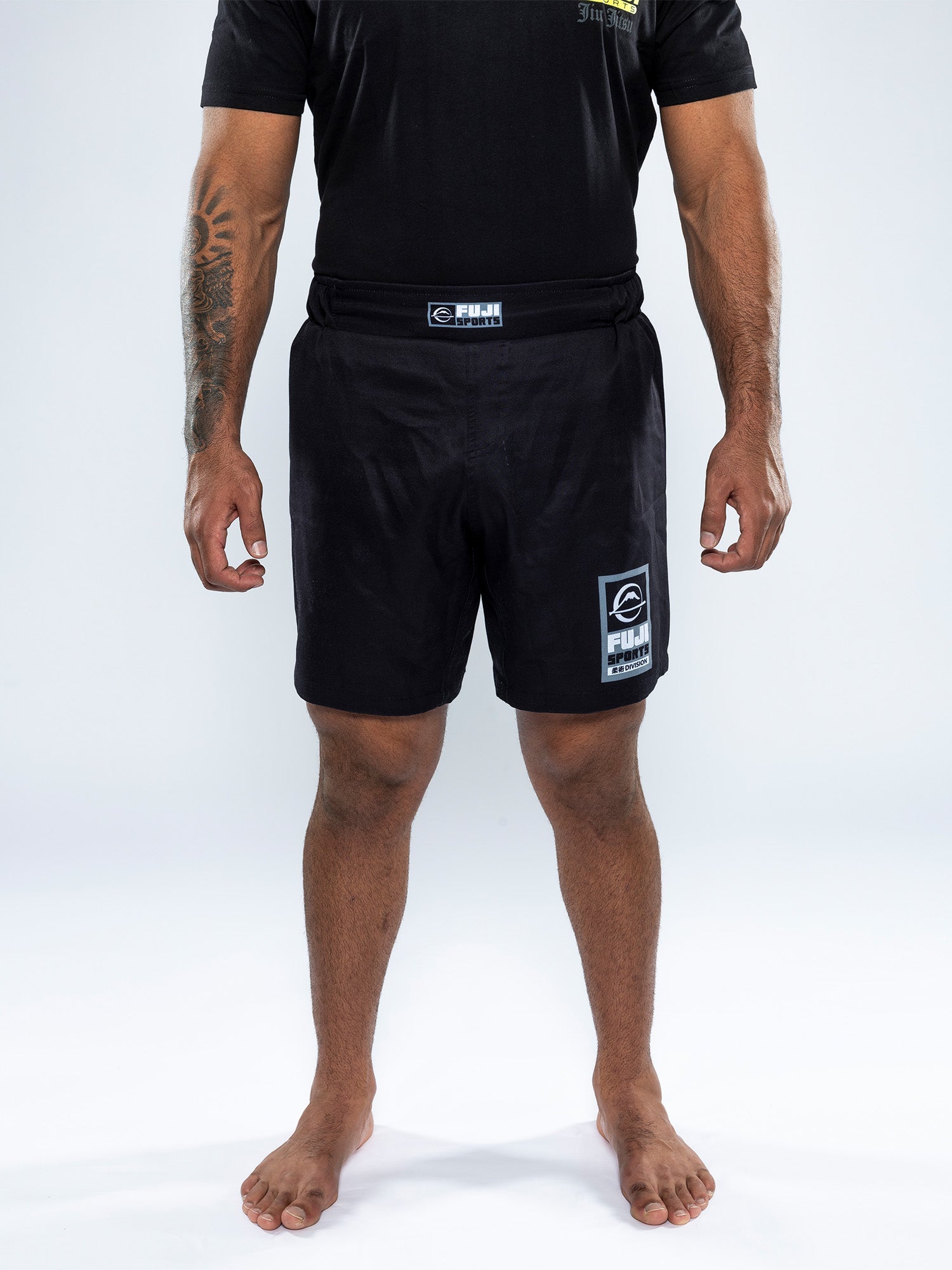 A person stands barefoot on a white background, wearing a black t-shirt and the Ultimate Grappling Shorts Black, which feature logos on the waistband and lower left leg. Their arms are relaxed at their sides.