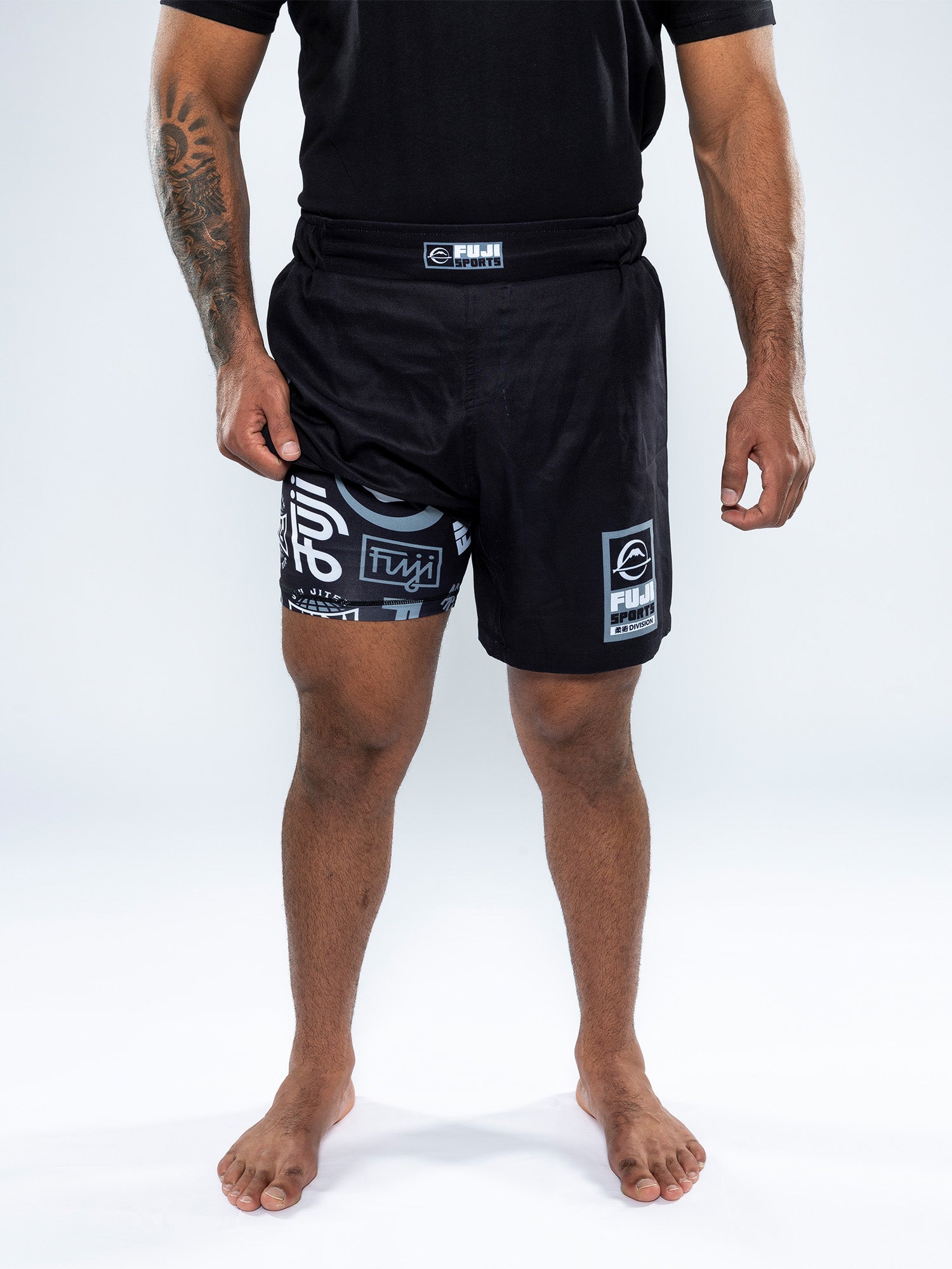 A person stands barefoot on a white background, wearing the Ultimate Grappling Shorts Black with various logos. Their hands rest at their sides, and only the body from neck to feet is visible.