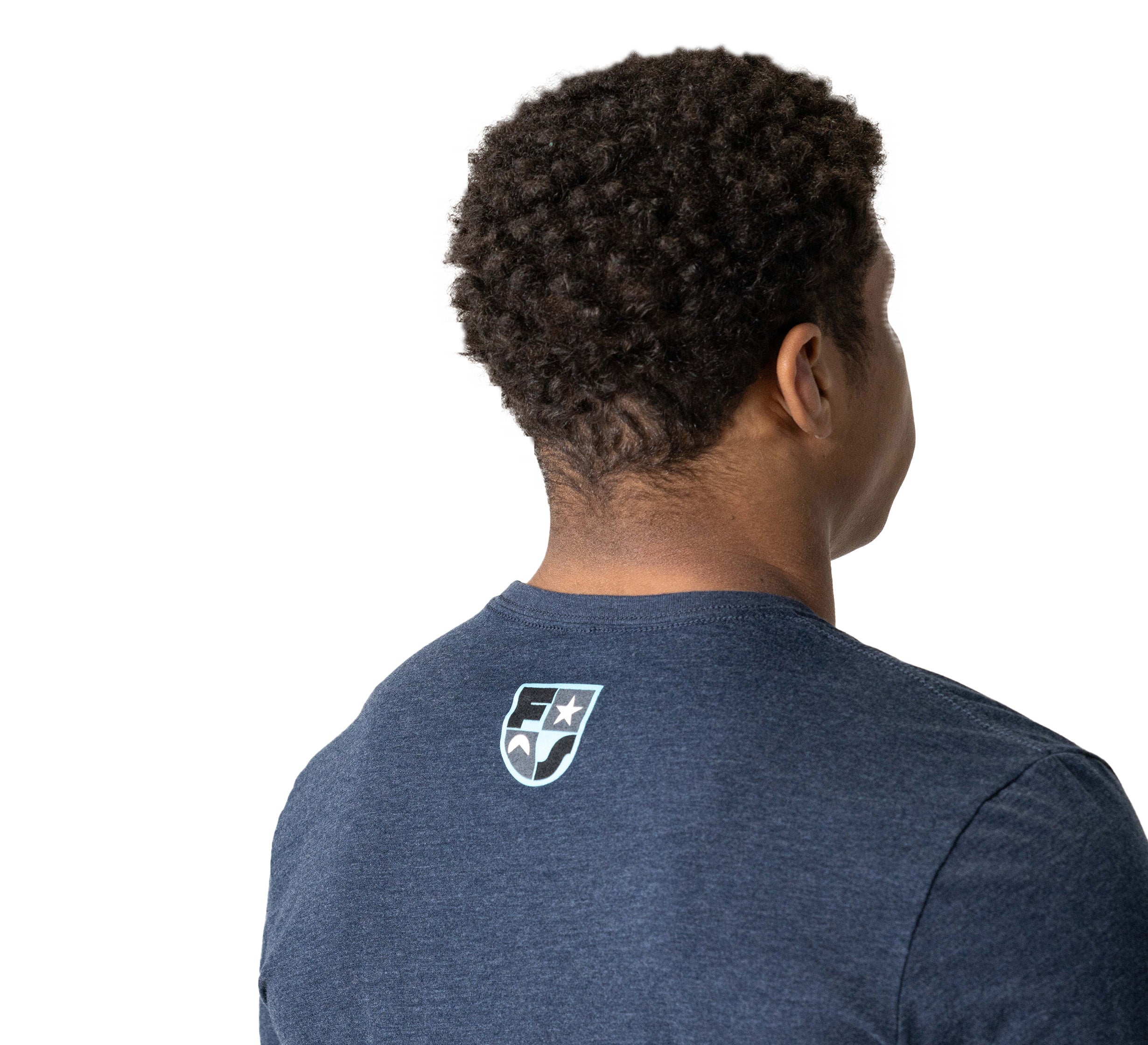 A person with short curly hair is shown from behind wearing the Retro Long Sleeve T-Shirt Navy, which features a small white shield logo on the upper back. The background is plain white.