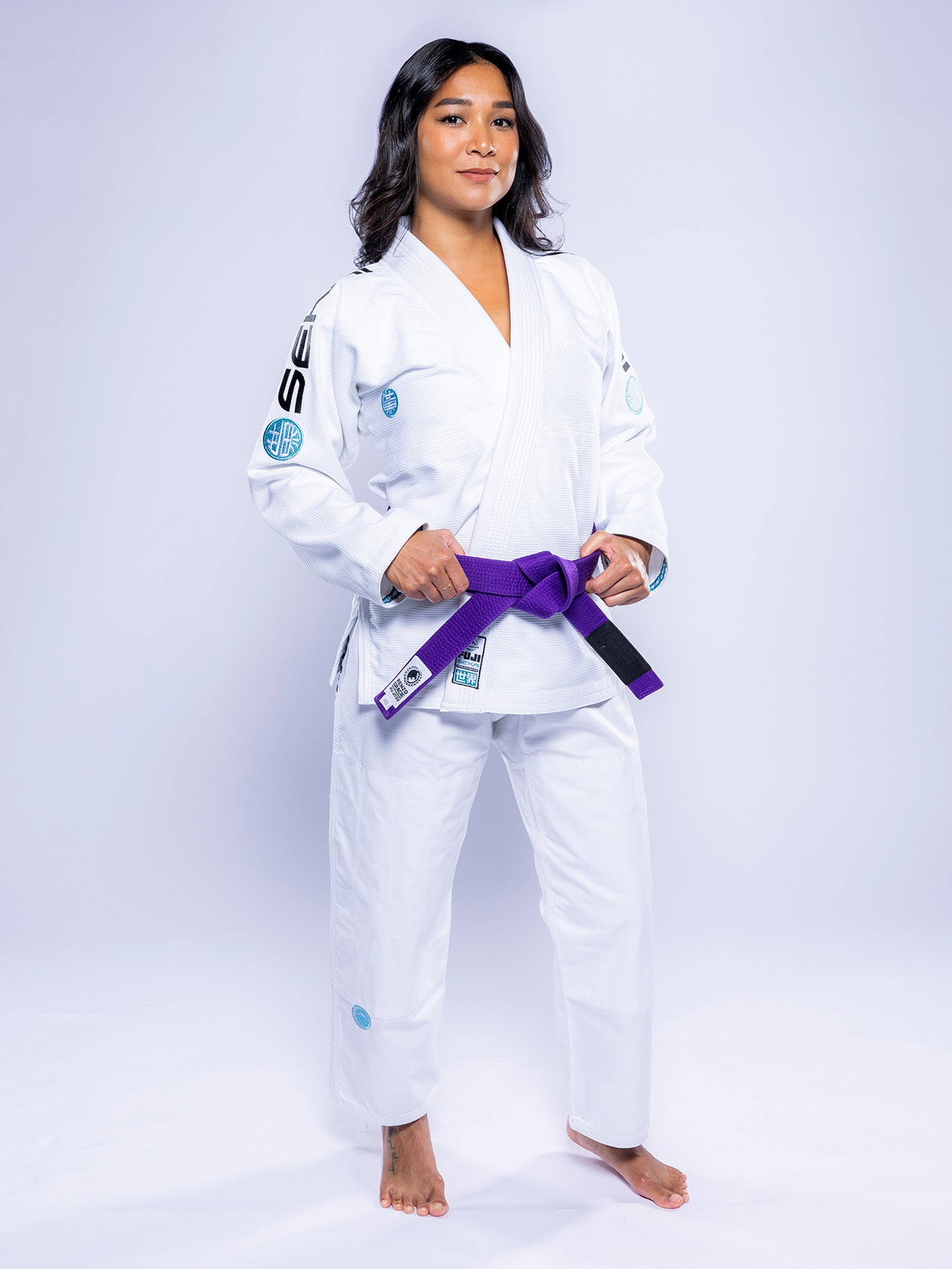 A woman stands barefoot on a white background, wearing the Womens SEKAI 3.0 BJJ Gi White Teal with a purple belt tied at her waist. She looks ahead confidently, hands resting on the belt.