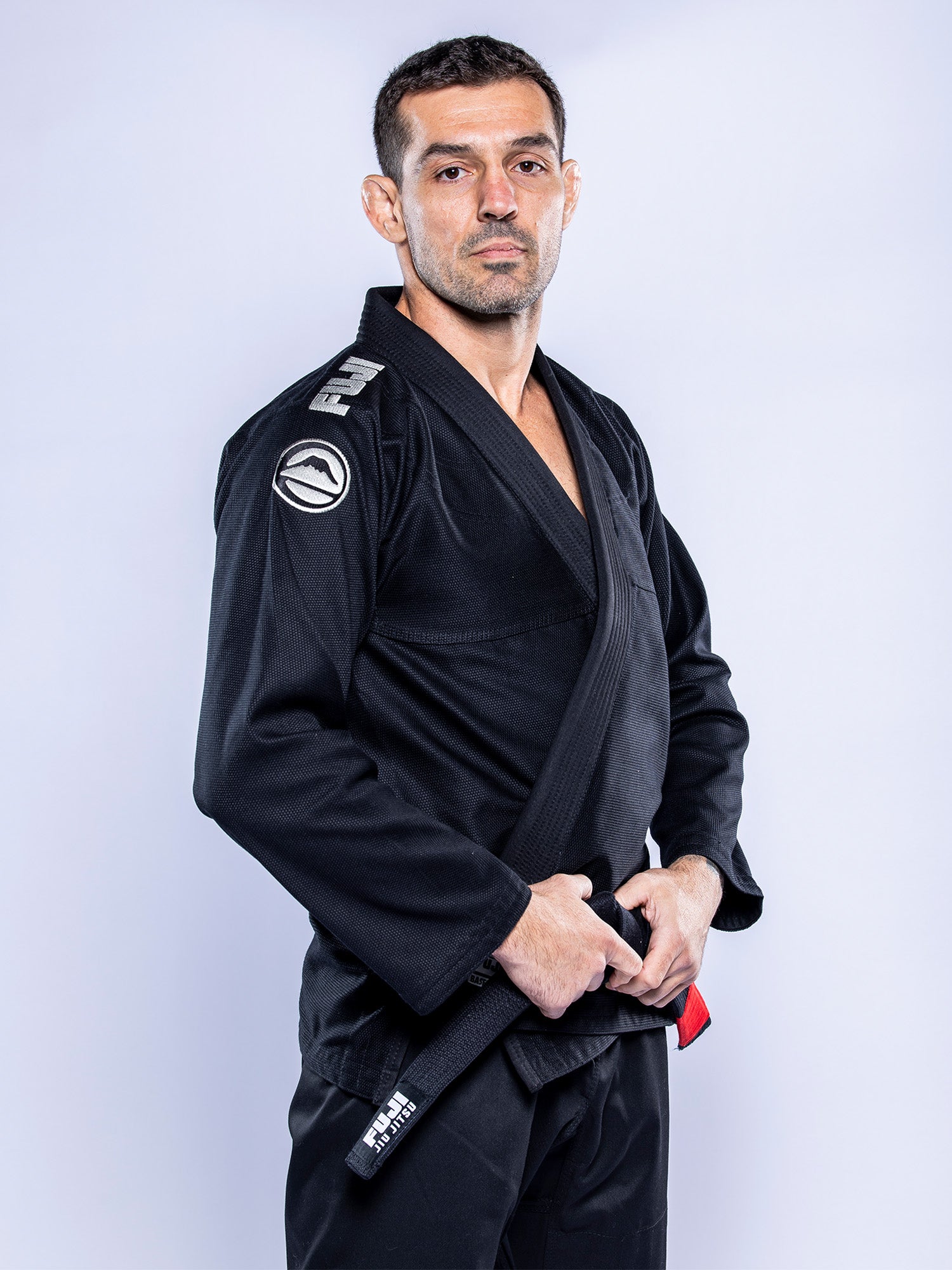 A man stands confidently against a plain light background, wearing the Baseline 2.0 BJJ Gi Black Grey and a black belt, with his hands resting near his belt as he looks at the camera.