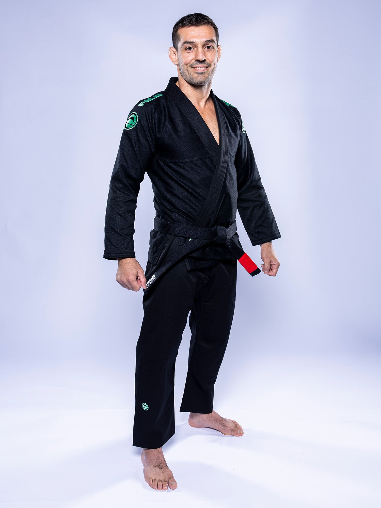 A man stands barefoot, smiling in the Baseline 2.0 BJJ Gi Black Mint with a black belt, set against a light blue studio background.