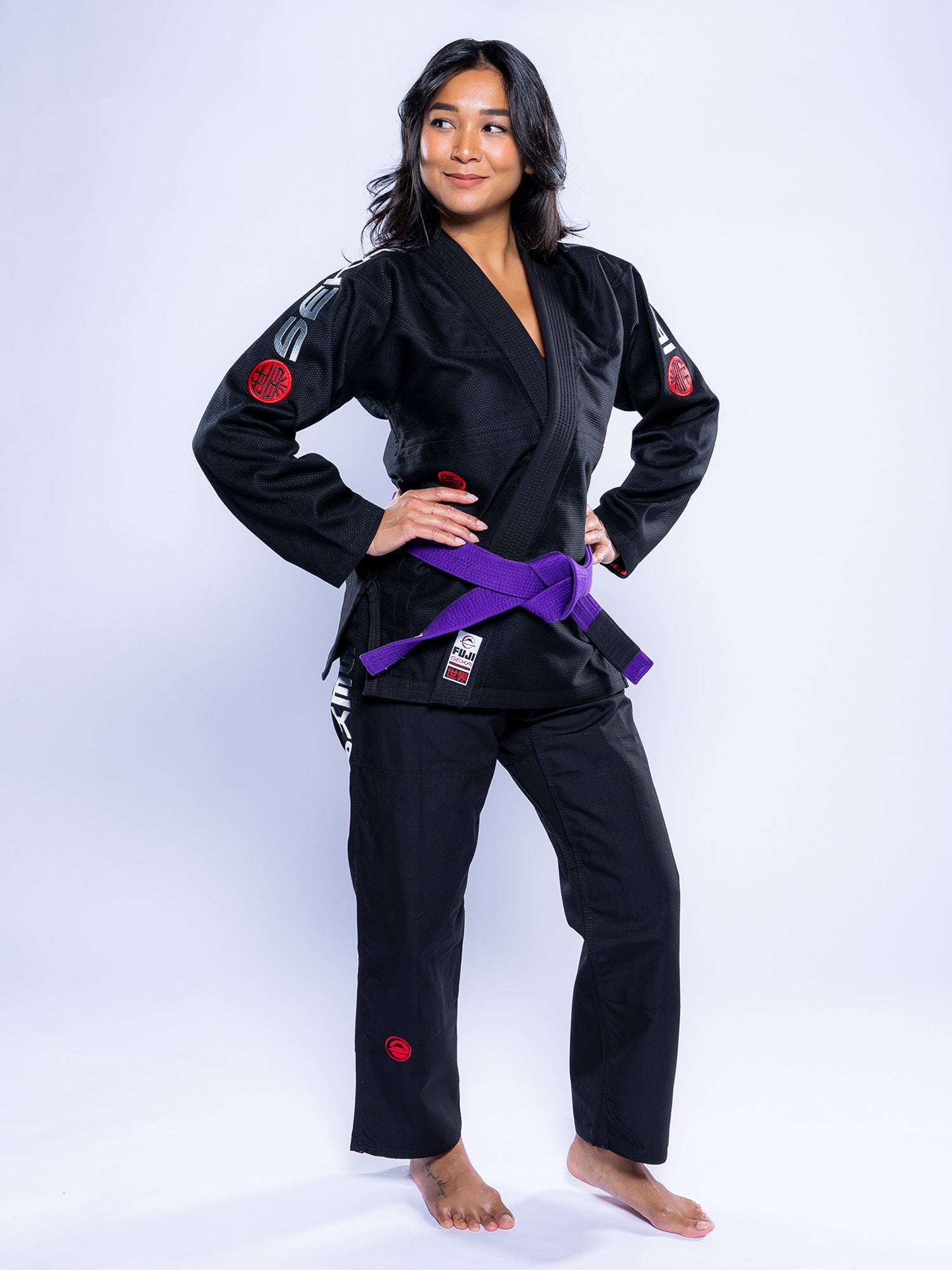 A woman stands barefoot on a white background, wearing the Womens SEKAI 3.0 BJJ Gi Black Grey with a purple belt. She has long dark hair, hands on hips, and looks confidently to the side.