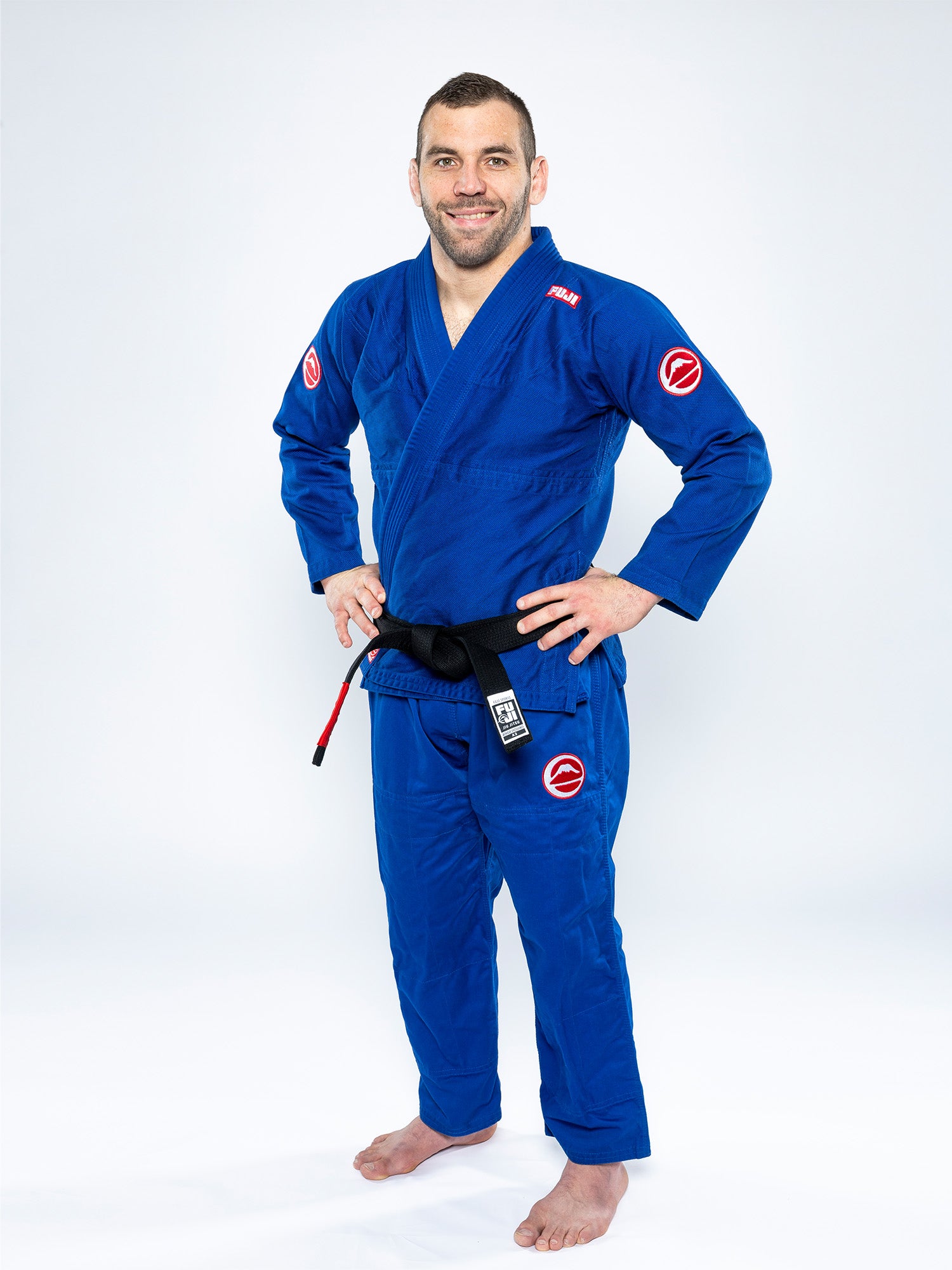 A man stands barefoot on a white background, smiling with his hands on his hips. He's wearing the Competition BJJ Gi Blue with a black belt, featuring visible patches on the sleeves and pants.