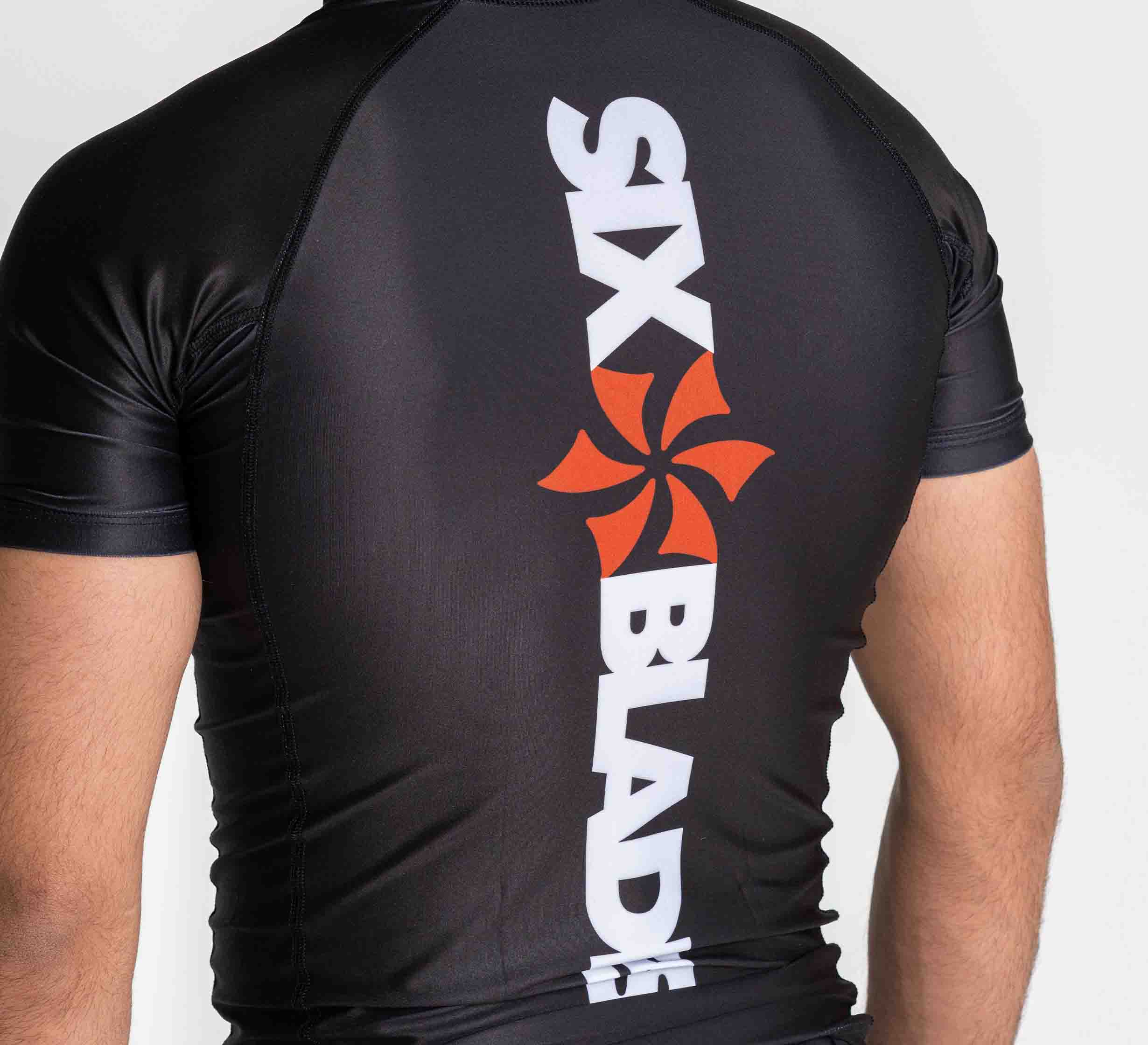 A person wears the Six Blades Short Sleeve Rashguard Black, featuring "SIX BLADE" in bold white letters and a red six-blade fan logo on the back. The snug fit highlights their upper back and arms.
