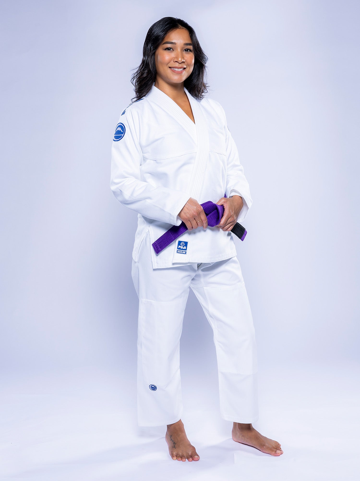 A person stands barefoot, smiling and wearing the Womens Baseline 2.0 BJJ Gi White Navy, holding a purple belt in front of them against a plain light background.