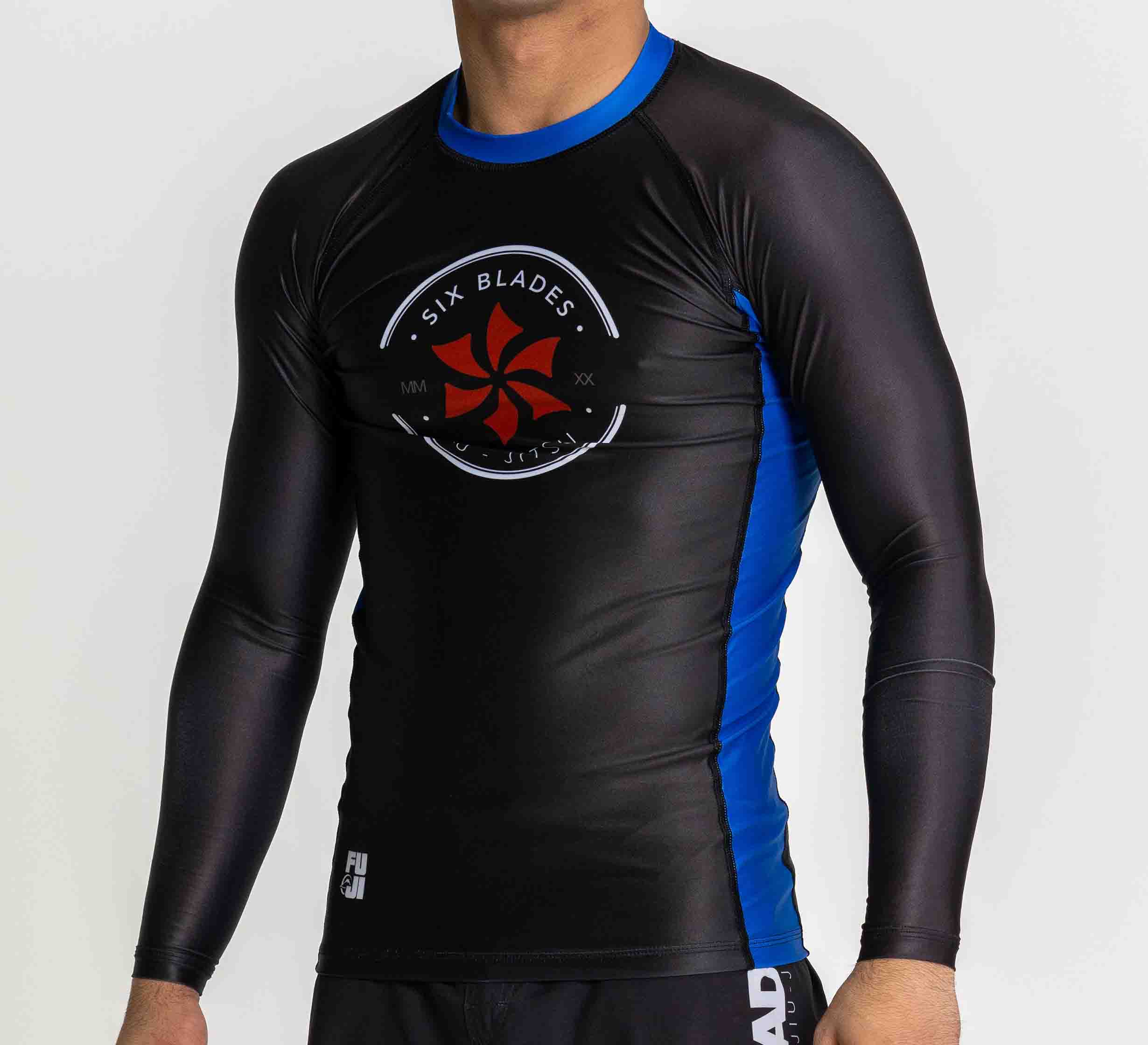 A person wearing the Six Blades Long Sleeve Rashguard Blue with a logo on the chest and black athletic shorts stands against a plain white background.