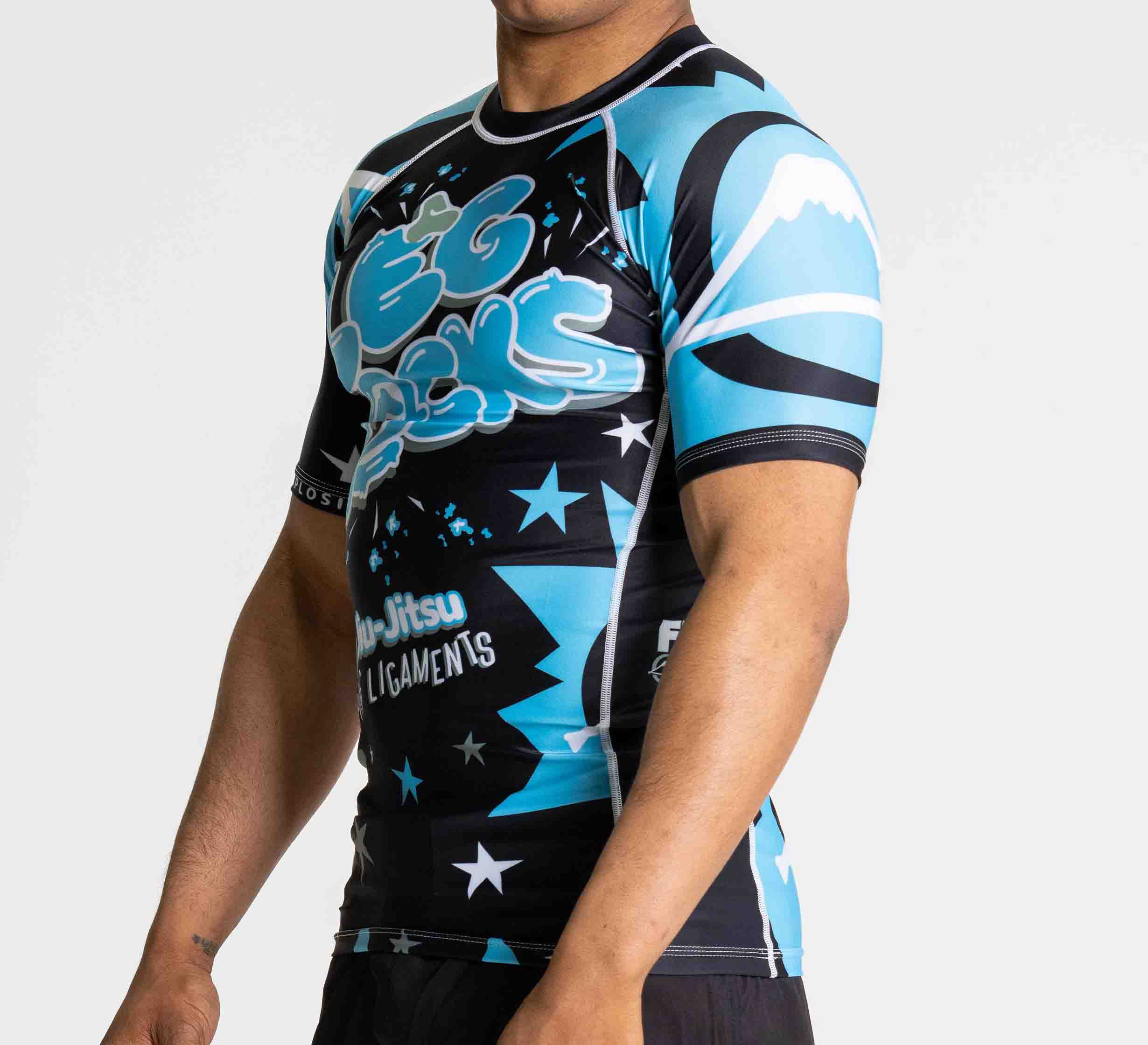 A person wearing the Leg Locks Flex Lite Rashguard Black, featuring blue, black, and white graffiti-style text, stars, and abstract designs, stands against a plain light background.
