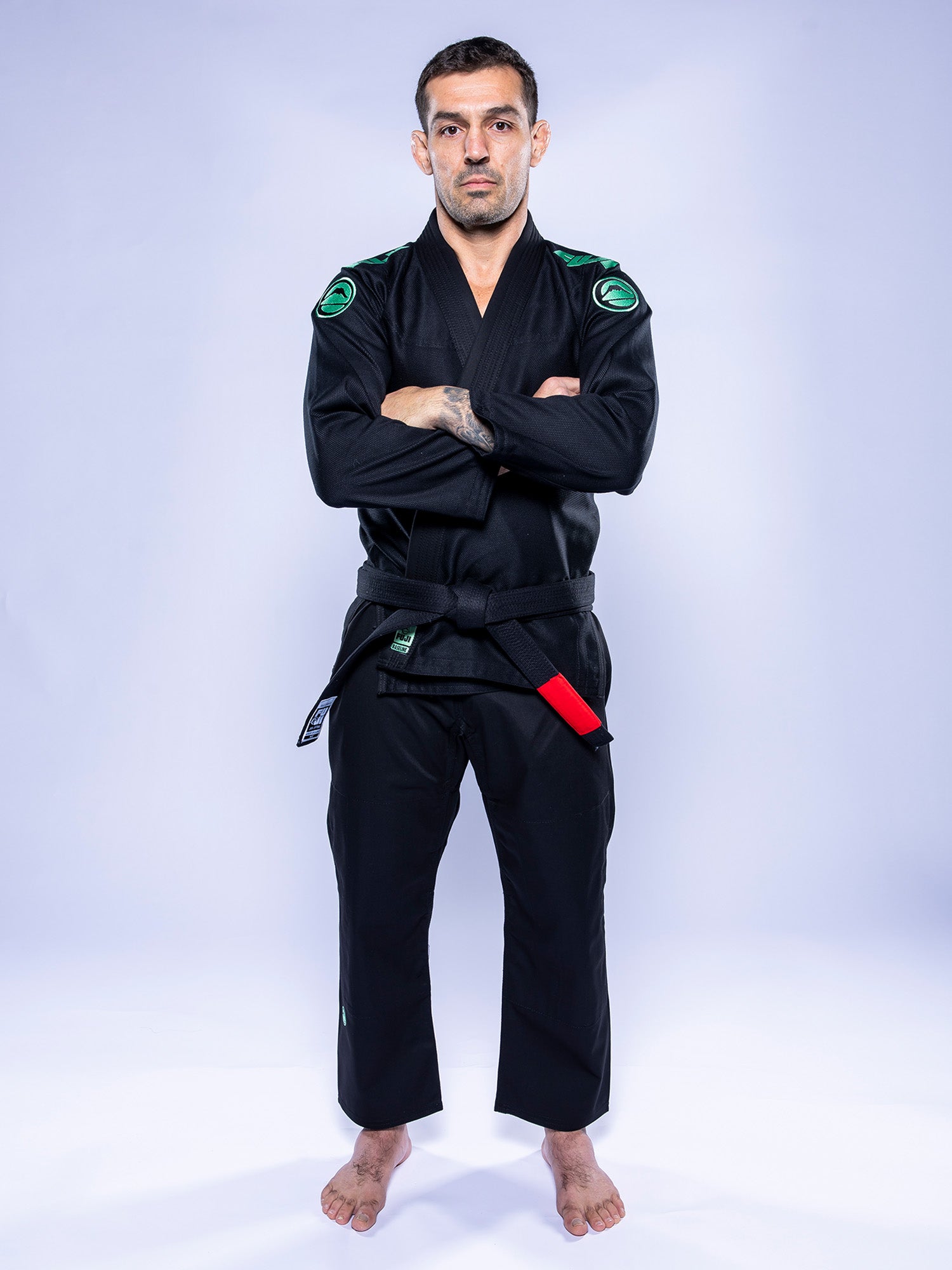 A man wearing the Baseline 2.0 BJJ Gi Black Mint with a black belt featuring a red bar stands barefoot on a white background, arms crossed and looking forward with a serious expression.