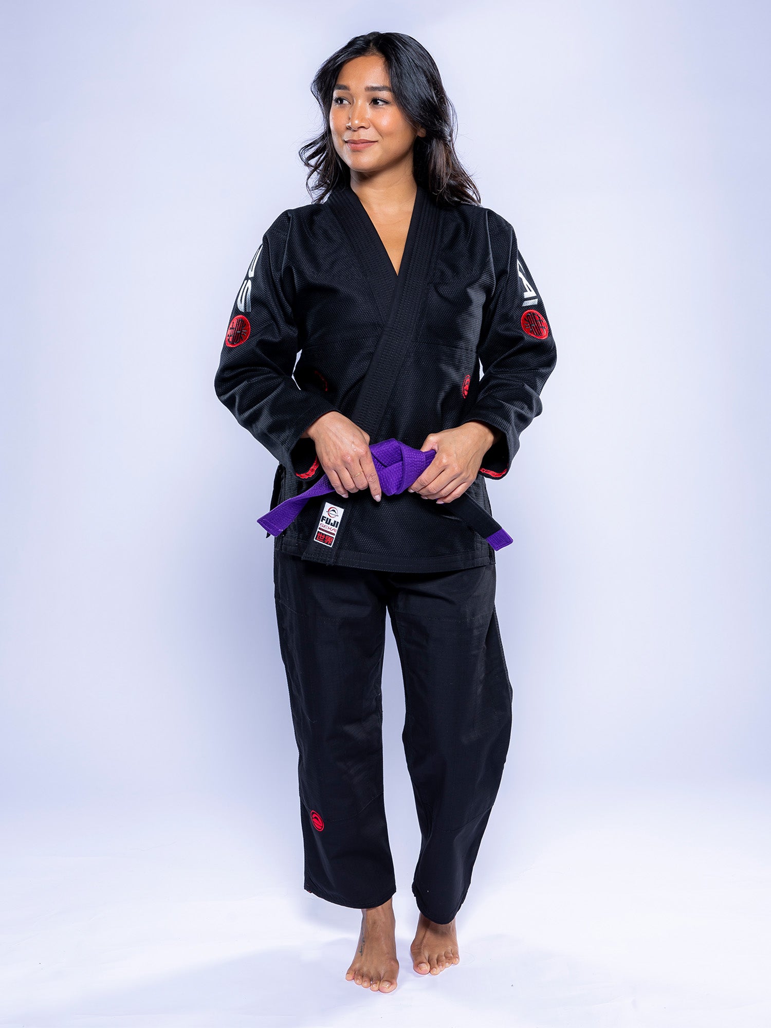 A woman in the Womens SEKAI 3.0 BJJ Gi Black Grey stands barefoot on a white background, holding a purple belt at her waist and looking to the side with a slight smile.