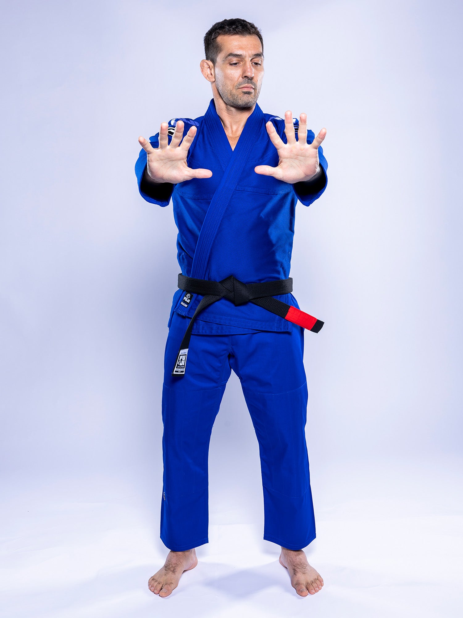 A man wears the Baseline 2.0 BJJ Gi Blue White with a black belt, standing barefoot on a white background, facing forward with arms extended and palms open in a defensive stance.