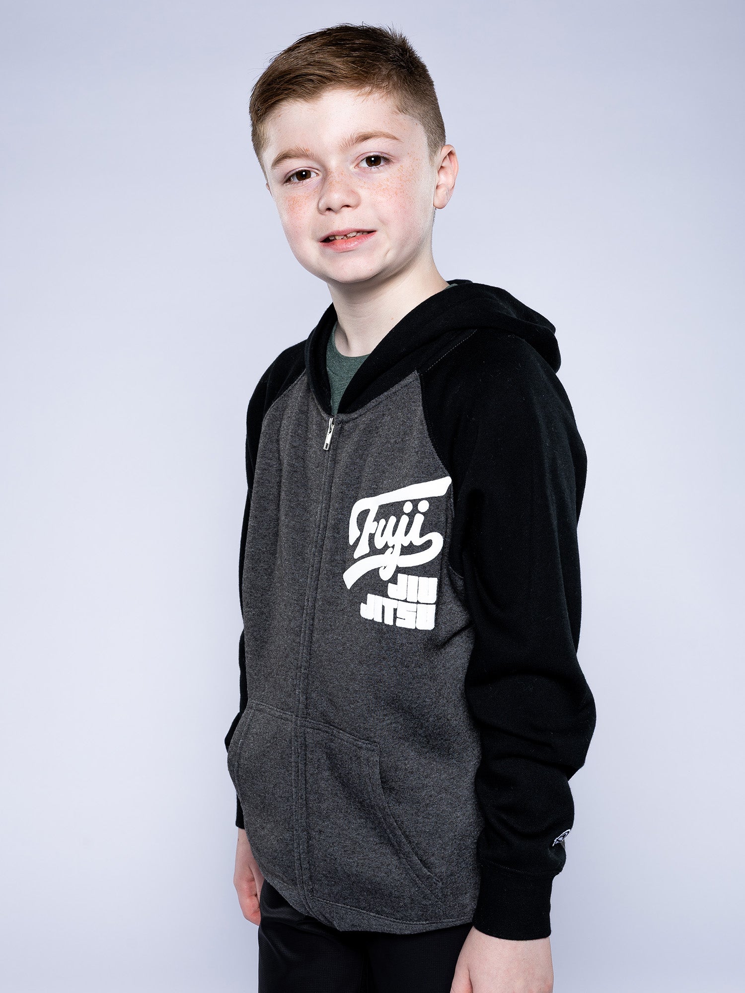 A young boy with short light brown hair smiles at the camera, wearing the Kids Jiu Jitsu Varsity Zip Hoodie in Charcoal, featuring "Fuji Jiu Jitsu" text on the chest, against a plain light gray background.