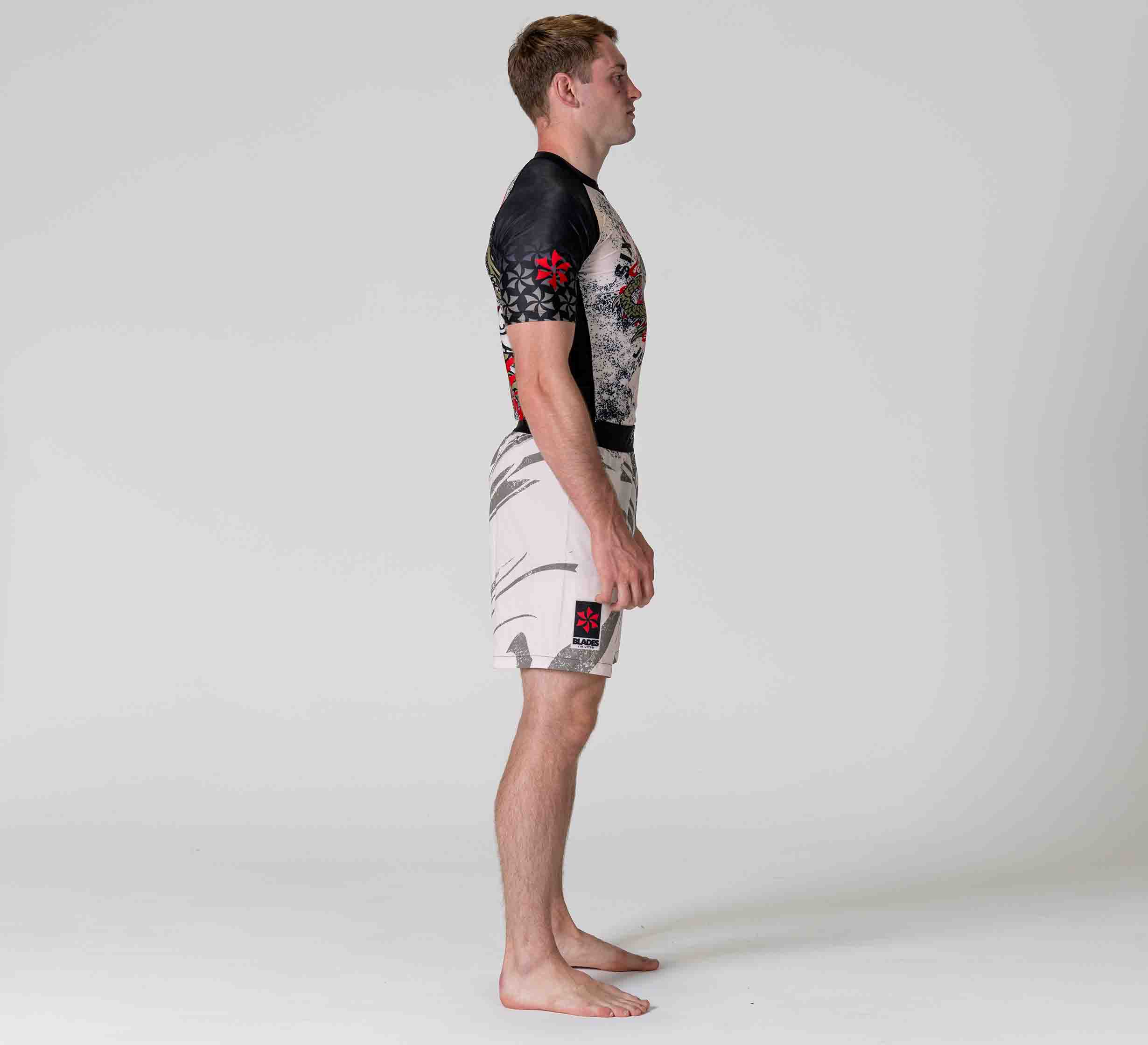 A man stands barefoot in profile against a plain background, wearing a black and grey athletic rash guard and the Six Blades Dragon Flex Lite Shorts, featuring graphic designs and red accents.