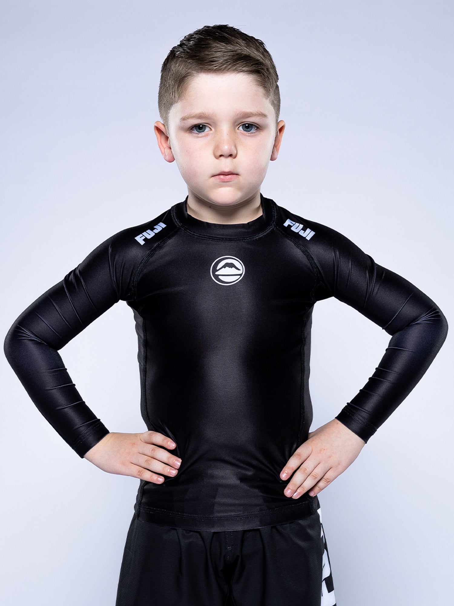 A young boy with short hair stands confidently with his hands on his hips, wearing the Kids Baseline 2.0 Long Sleeve Rashguard Black and matching black shorts against a plain light background.
